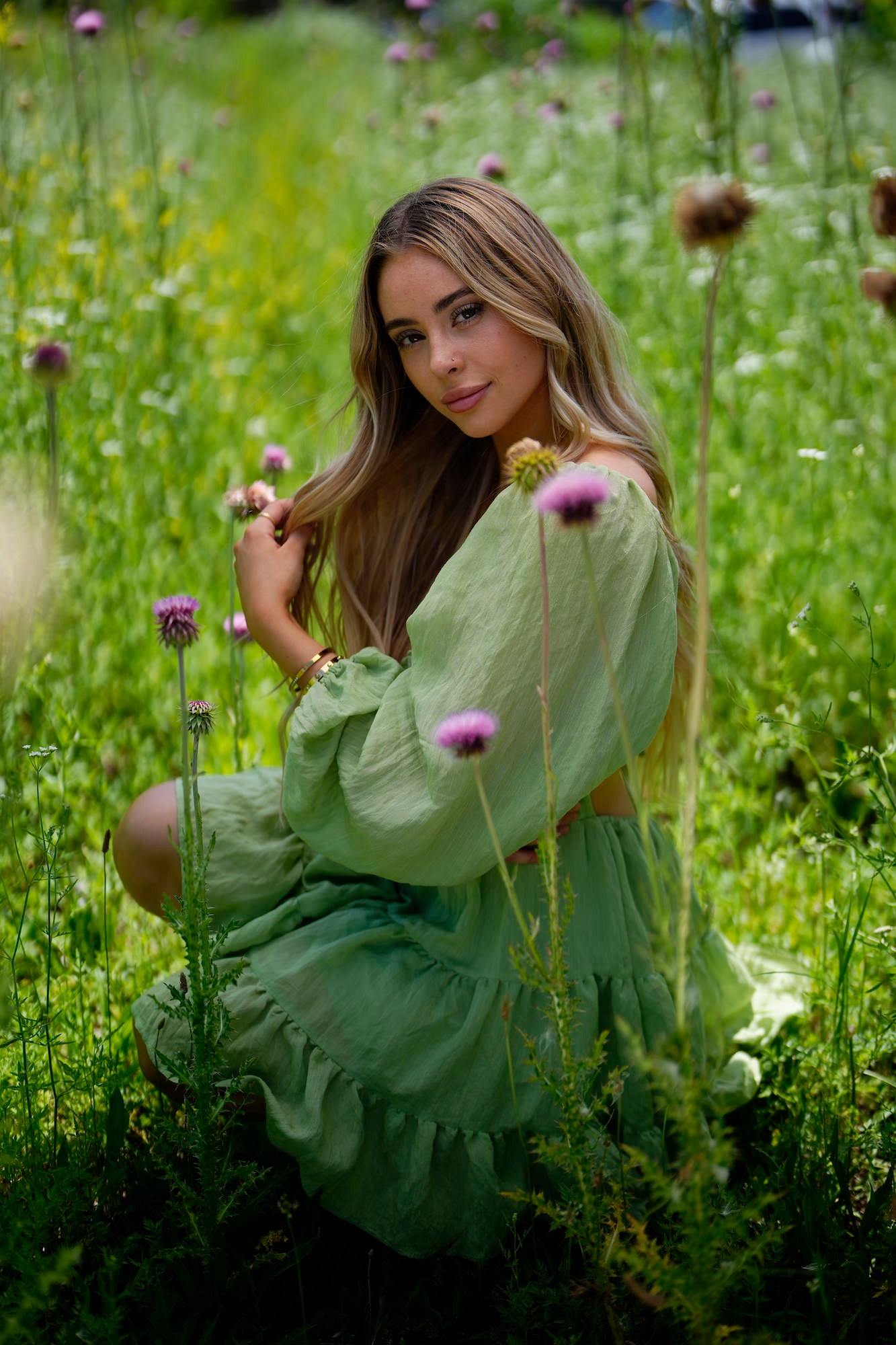 Woman in a green off-shoulder dress kneeling in a field of purple thistles and tall grass.