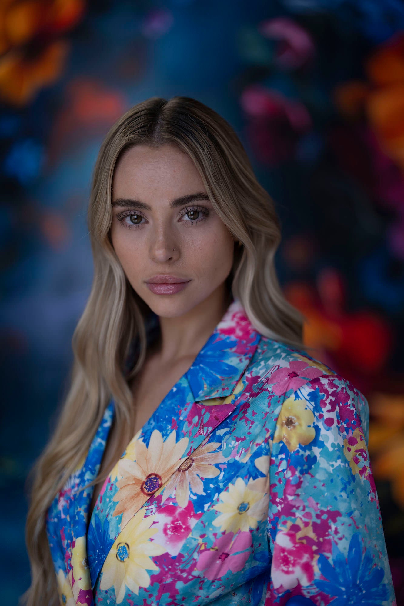 Blonde woman in a bright floral blazer looks at the camera against a blurred colorful floral backdrop.