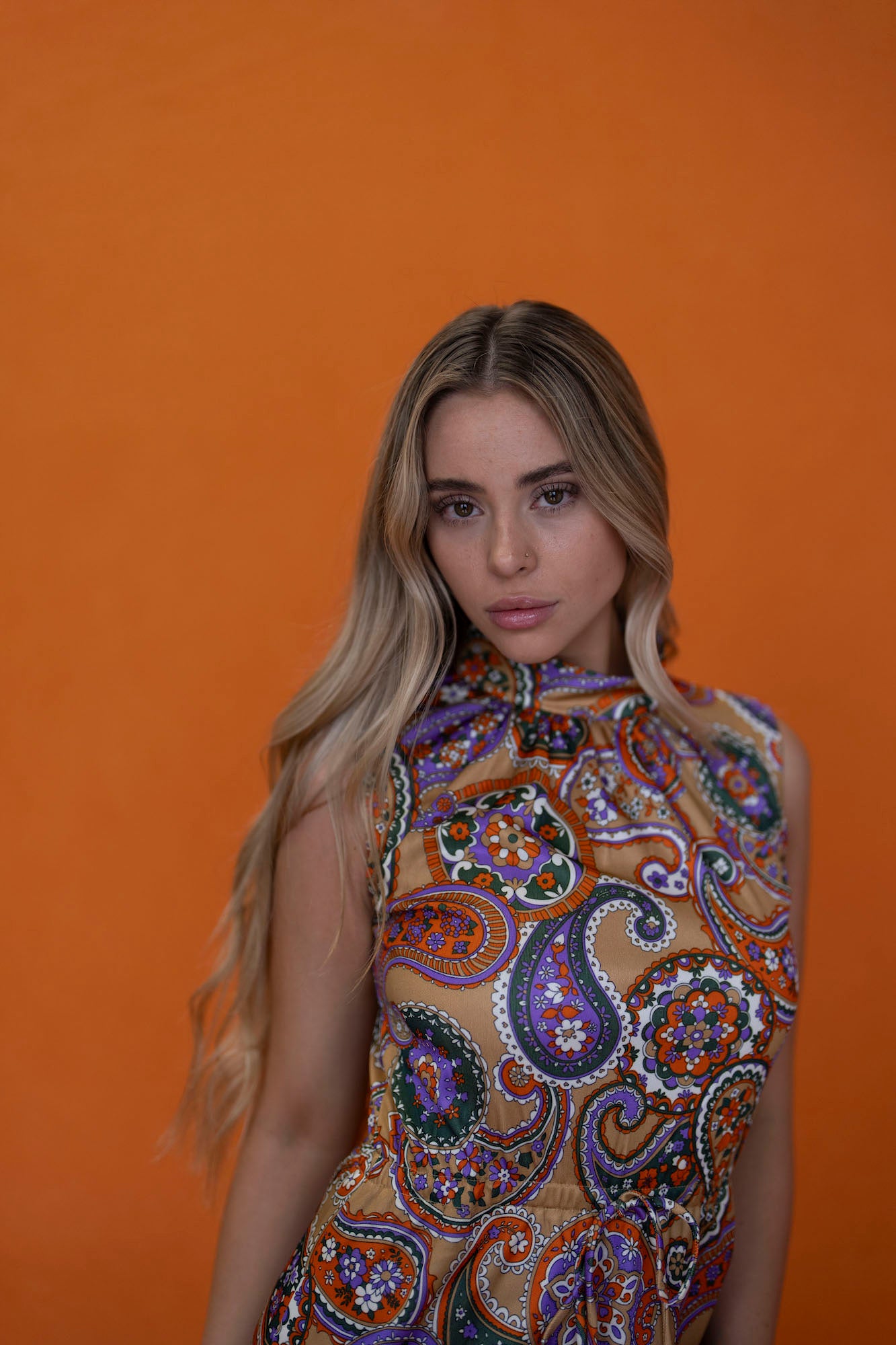 Blonde woman in a paisley-print sleeveless top posing against a bright orange wall.