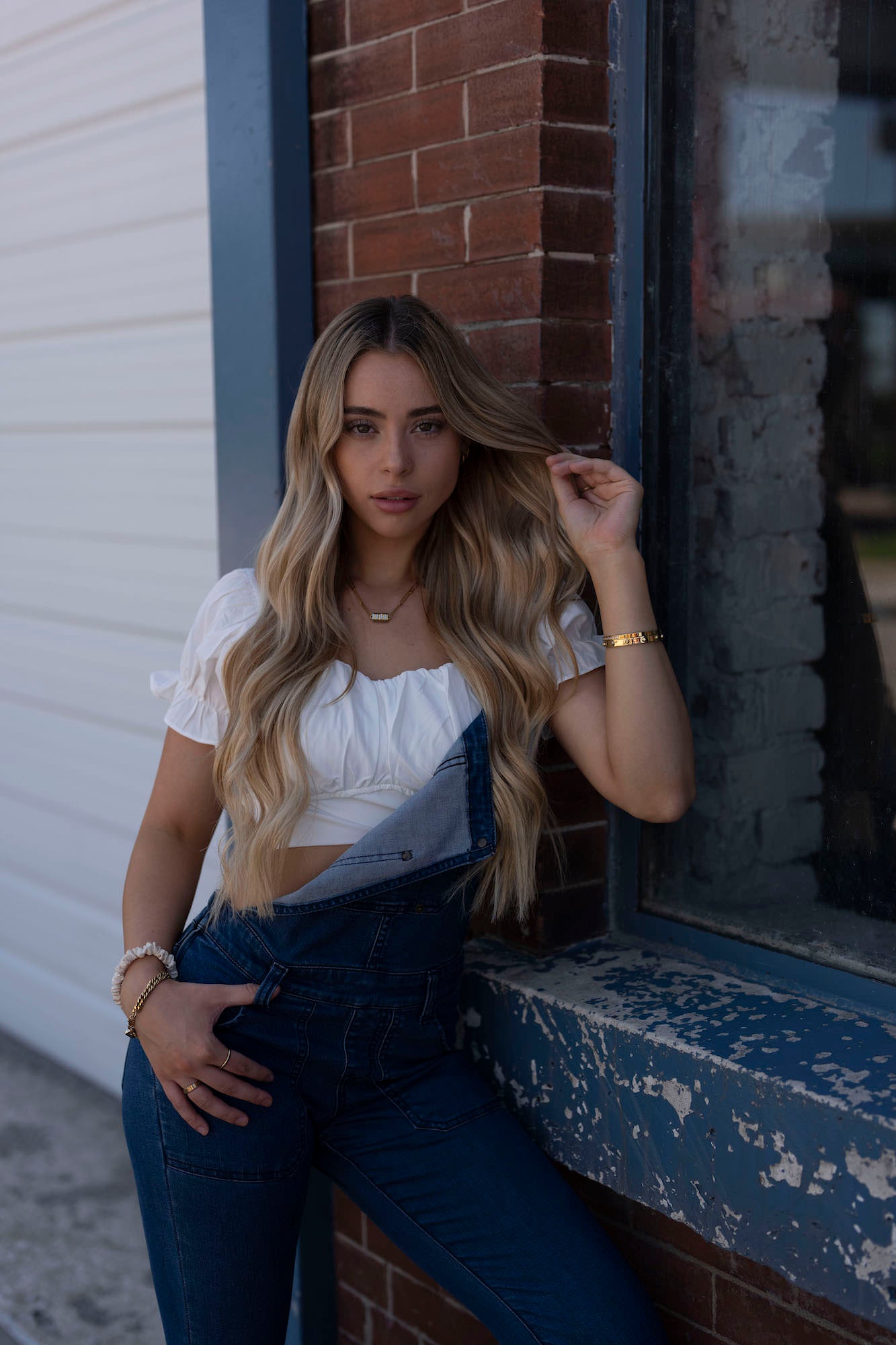 Woman with long blonde hair in denim overalls and white top leaning against a brick wall by a window.