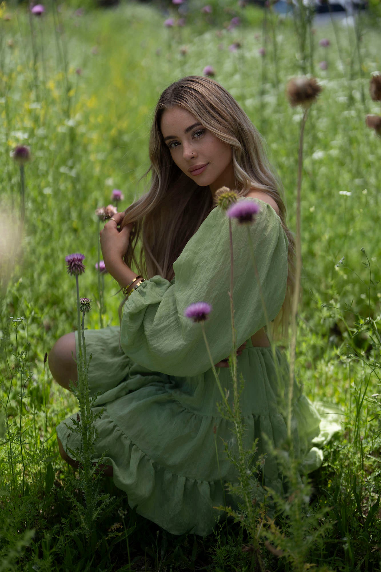Woman in a green off-shoulder dress kneeling in a meadow of purple thistles and tall grass.