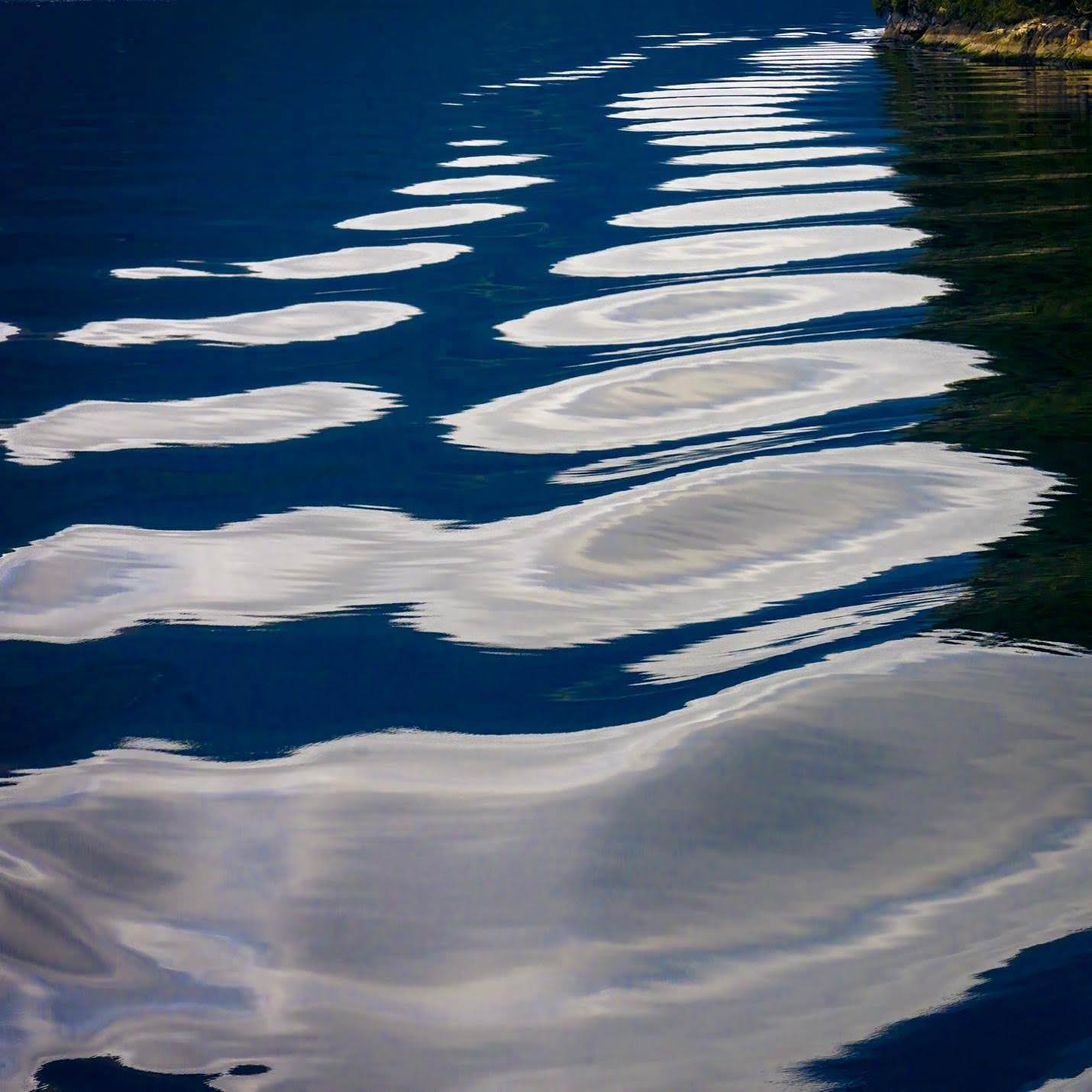 An abstract close-up of rippling water reflects cloud formations in soft white ovals across a deep cobalt surface, revealing the quiet artistry hidden in New Zealand's fiords, as seen by Don Smith.