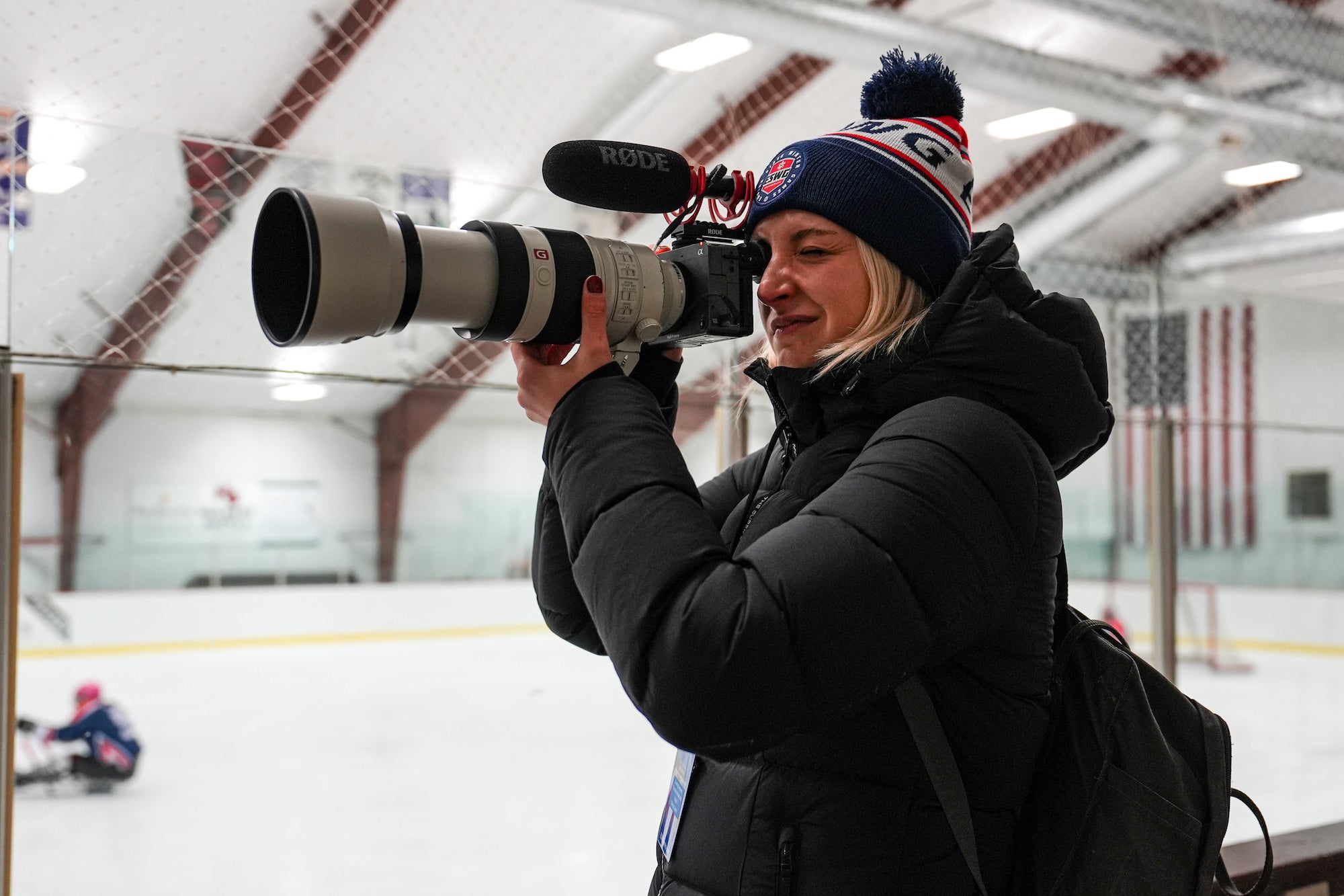 Videographer in black parka films through glass with telephoto and mic on rink.
