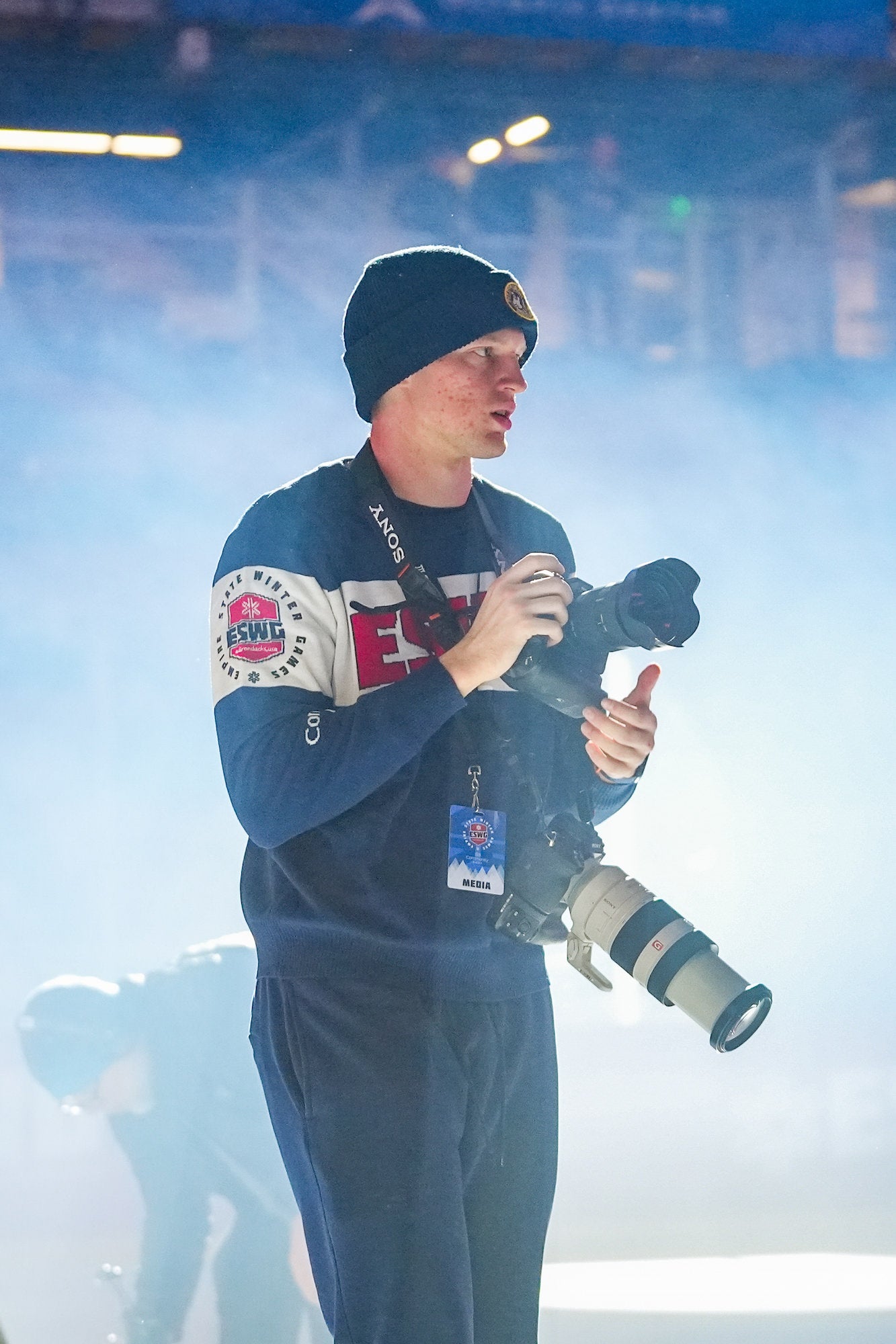 Photographer with media badge holds camera and telephoto lens amid rink fog.