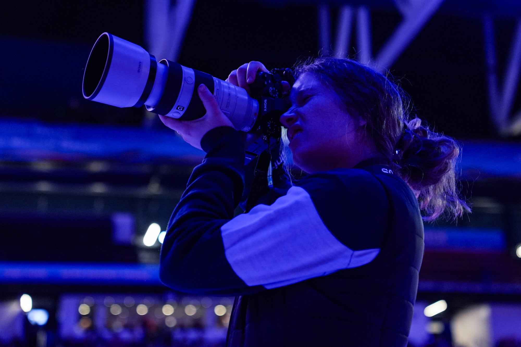 Photographer with long telephoto lens shoots from stands under purple arena lights.