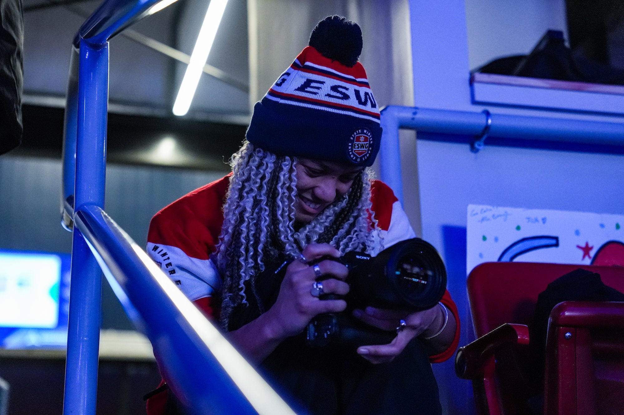 Photographer in ESWG beanie reviews camera on arena steps under blue light.