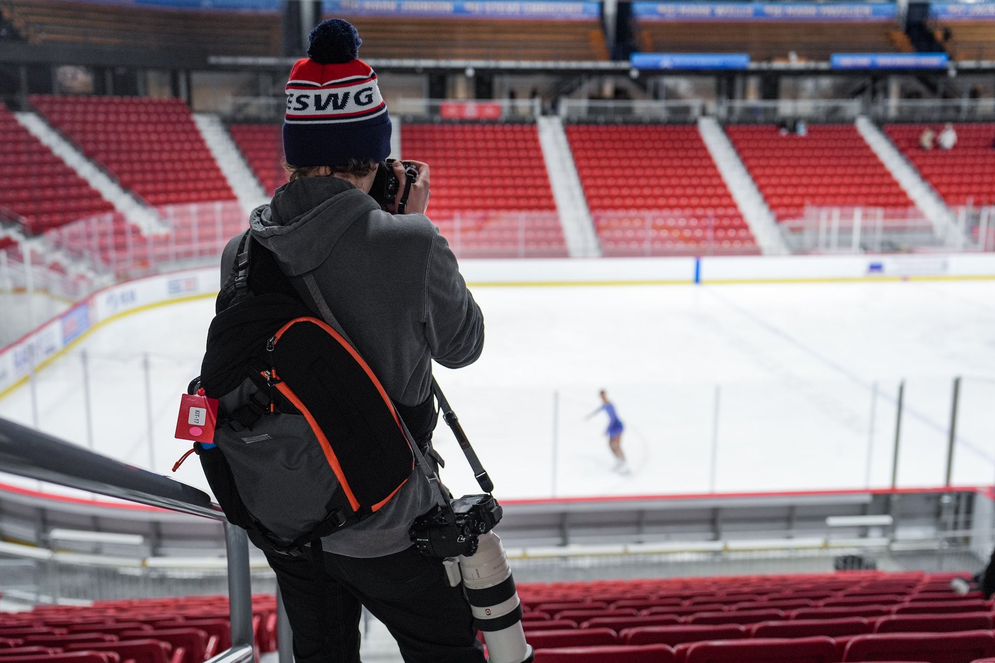 Photographer in stands aims camera at lone figure skater on rink below.