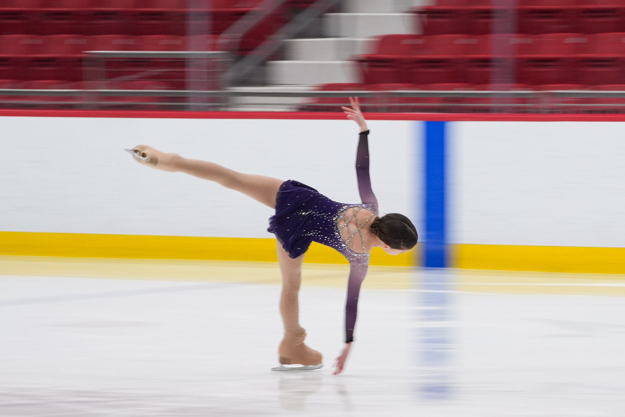 Figure skater in a purple dress executes a deep spiral with one leg extended back; motion blur and red arena seats in background.