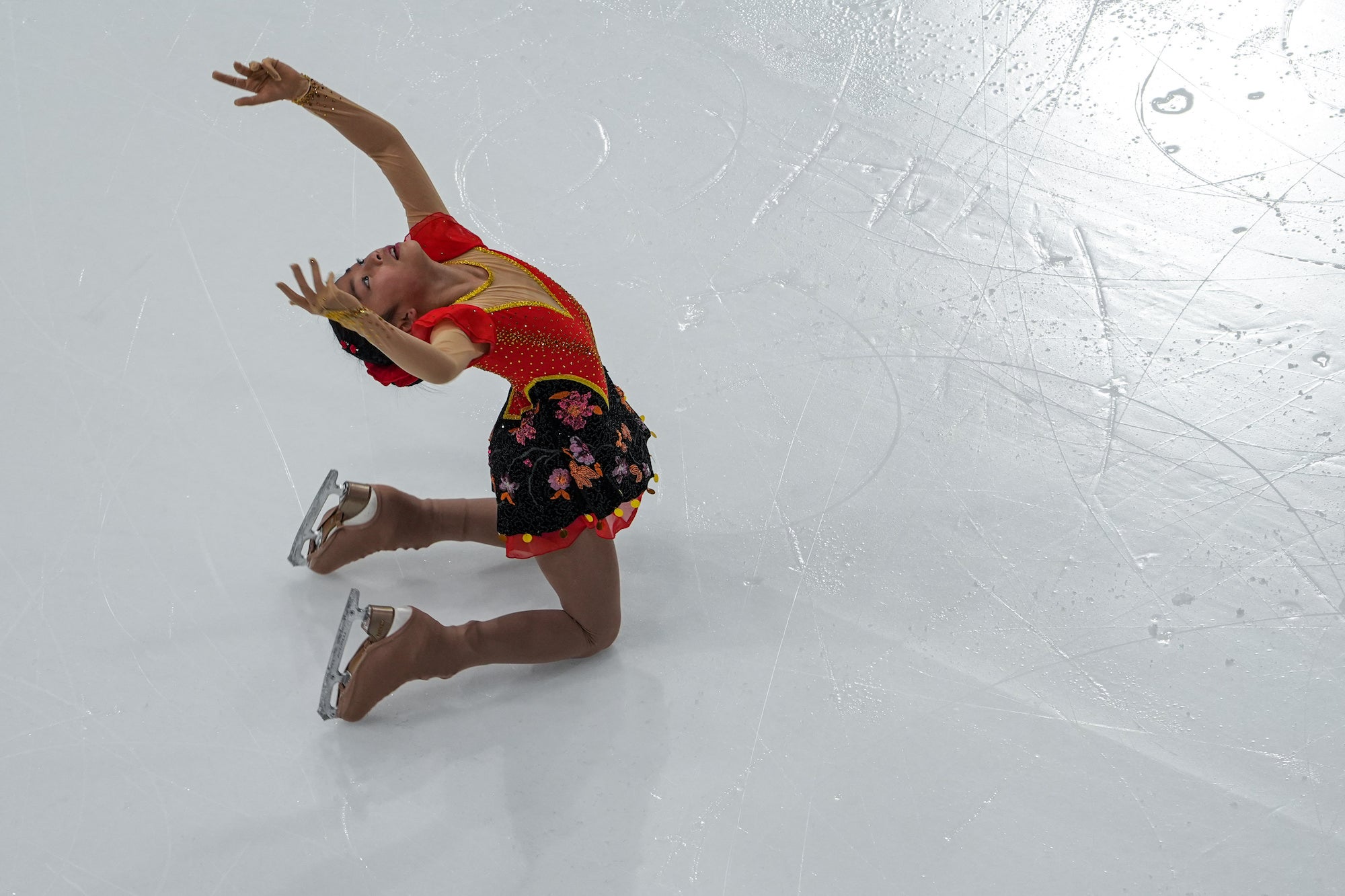 Figure skater in a red and black floral dress arches backward with raised arms during a routine on bright, marked ice.