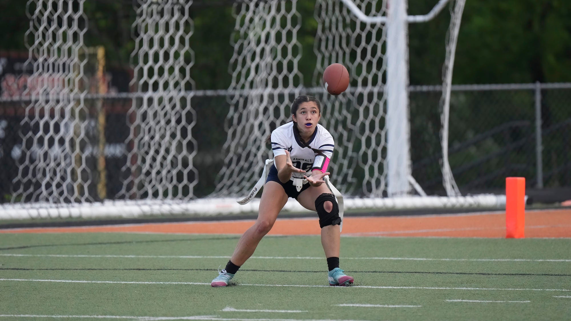 A female flag football player in a white Admirals jersey crouches low on a turf field, hands extended to receive a snap, with a football in mid-air above her and an orange end zone pylon visible in the background.