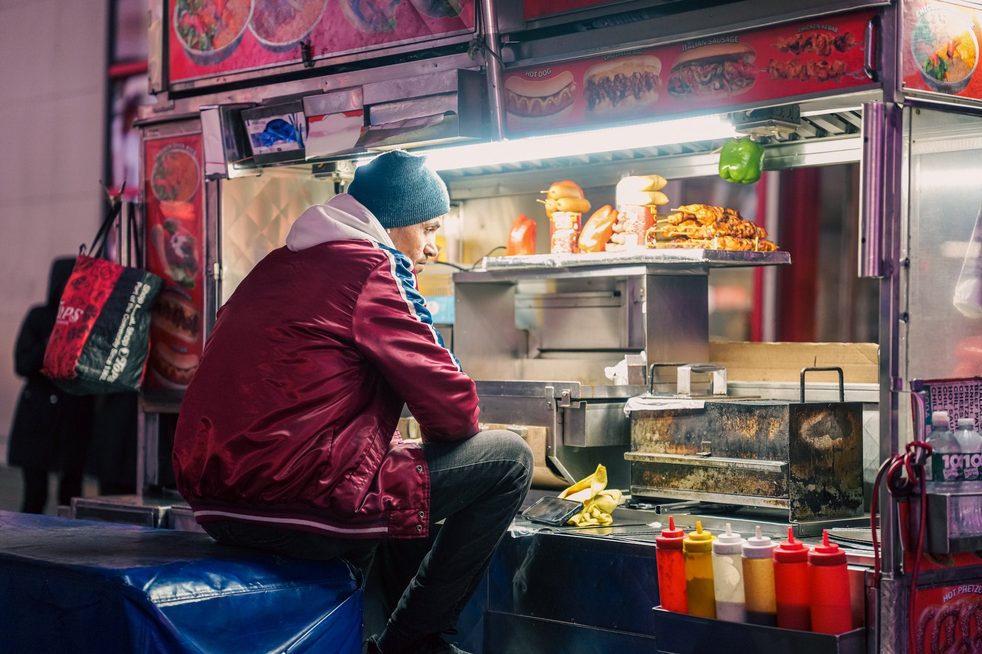 A street food vendor wearing a red jacket and blue beanie sits perched on a stool at his illuminated cart at night, with hot dogs and sausages displayed on a shelf, and condiment bottles lined up in front of him.