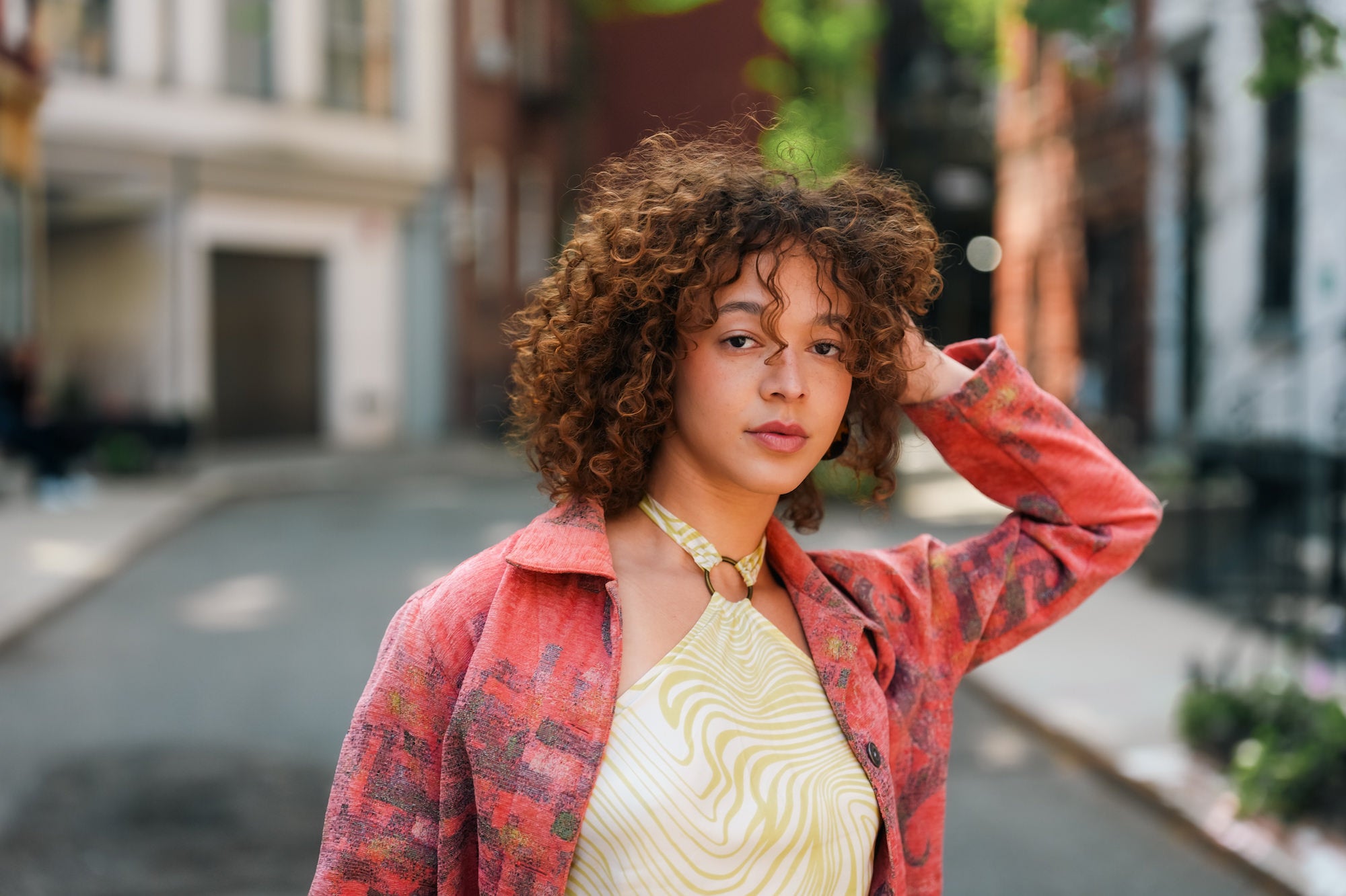 Environmental portrait of a young woman with curly hair standing on a city street, wearing a red patterned jacket over a yellow swirl-print top, one hand raised to her hair, with blurred urban buildings behind her.