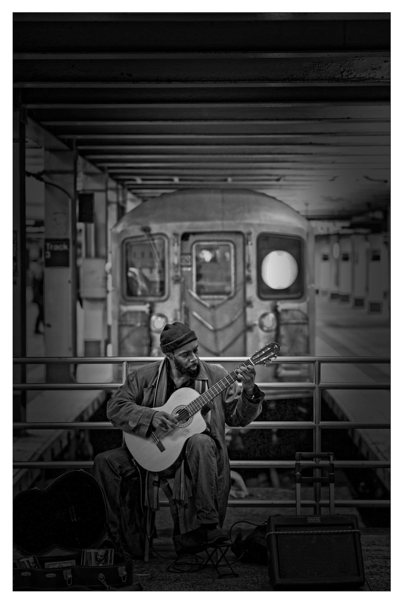 Subway platform musician plays classical guitar as a train approaches in the background; monochrome.