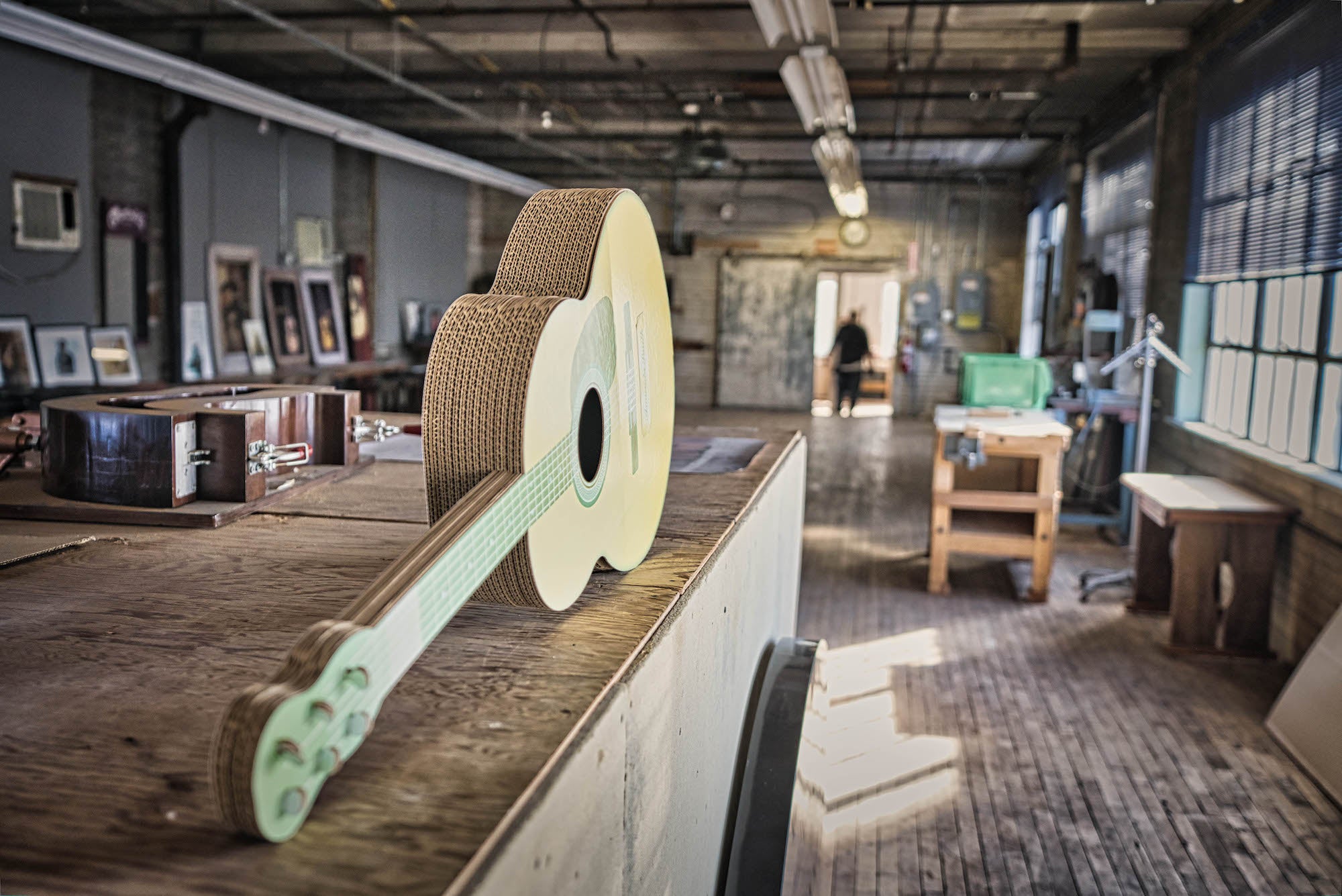 Workshop interior with a cardboard mockup guitar on a long bench; tools and frames line the room.