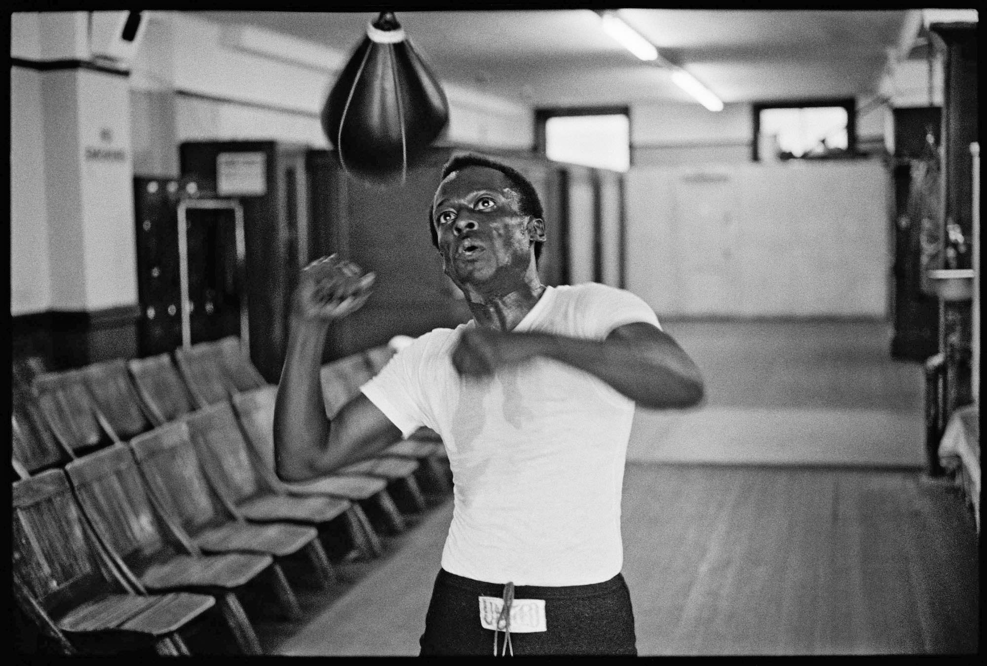 Sweat‑drenched boxer works a speed bag in an empty gym corridor, chairs lined along the wall.