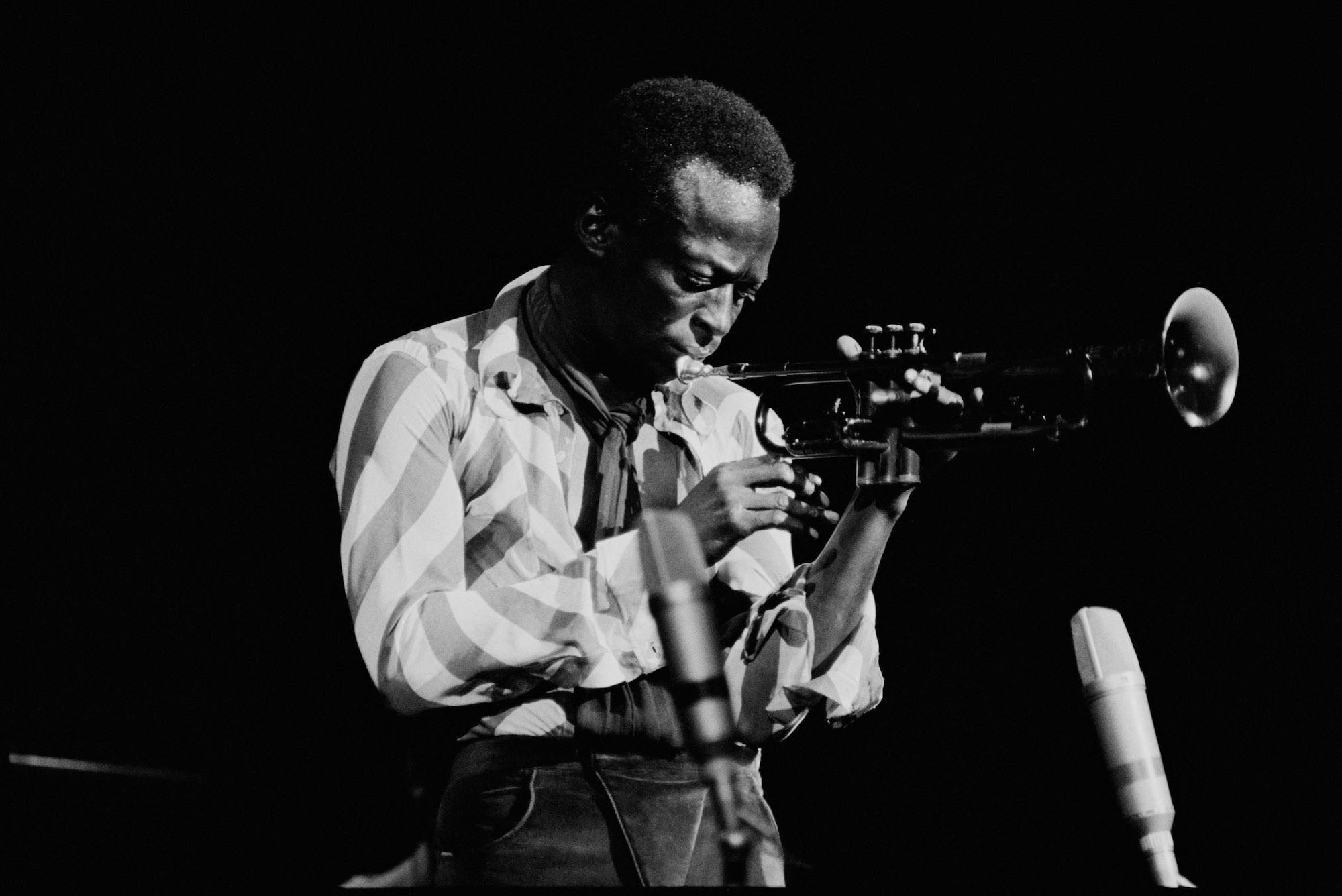 Jazz trumpeter in a striped shirt plays under a spotlight on a dark stage; microphones frame the scene.