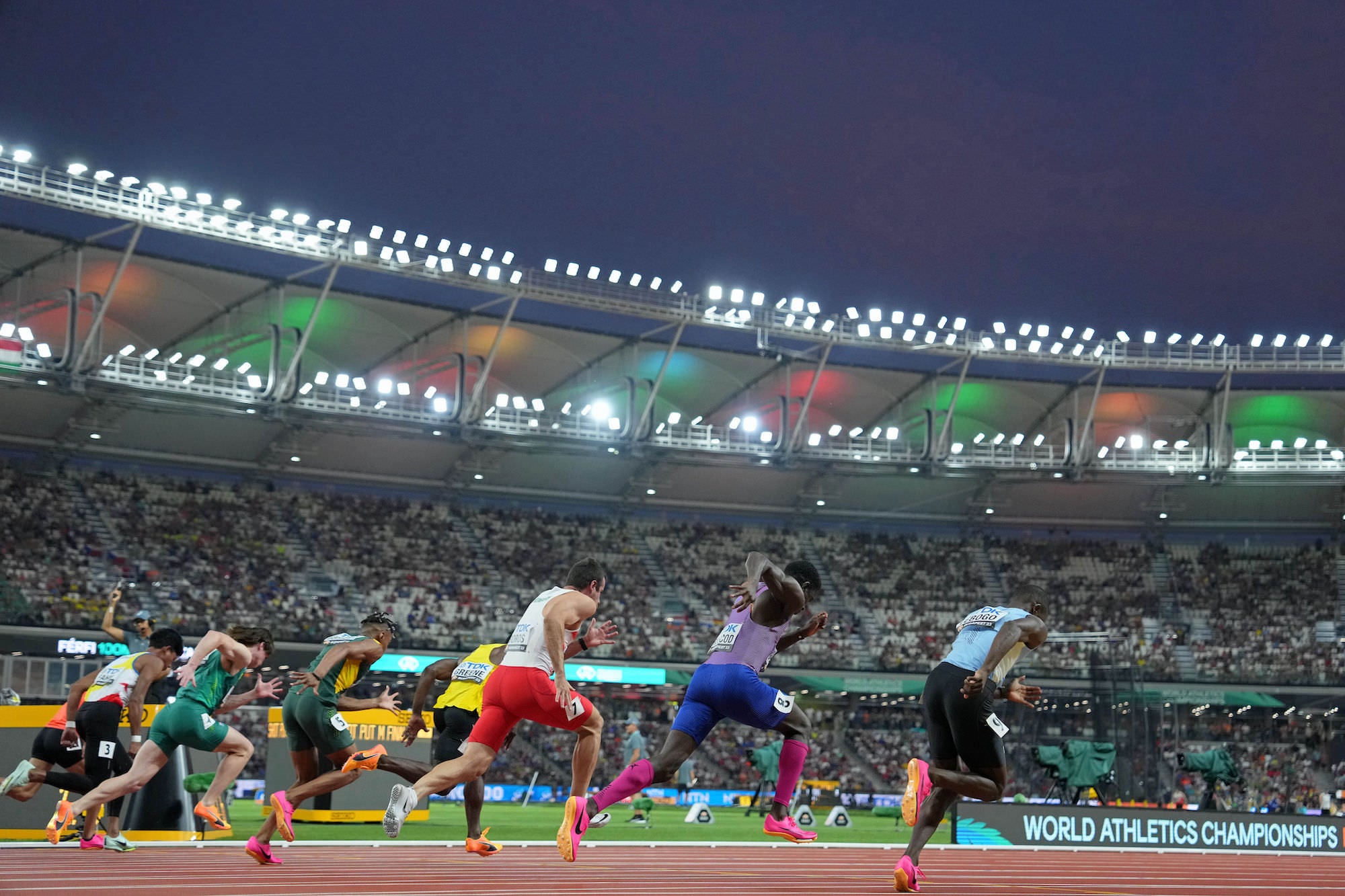 Sprinters explode from the blocks in a packed stadium at dusk. Sprinters explode from the blocks in a packed stadium at dusk.