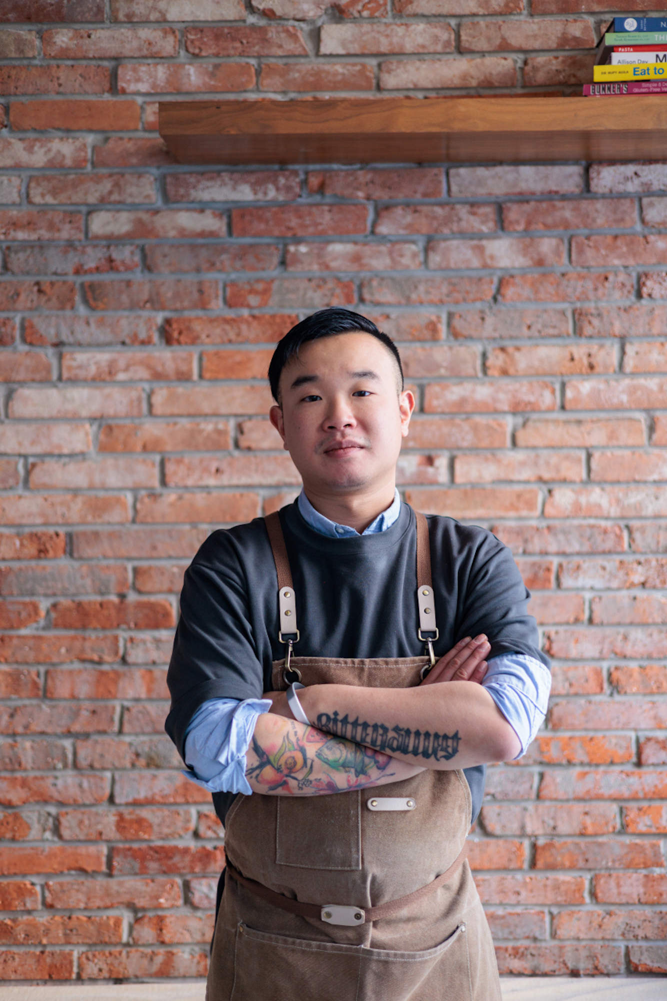 Isaac Tang in apron stands with arms crossed against a brick wall, tattooed forearms visible.