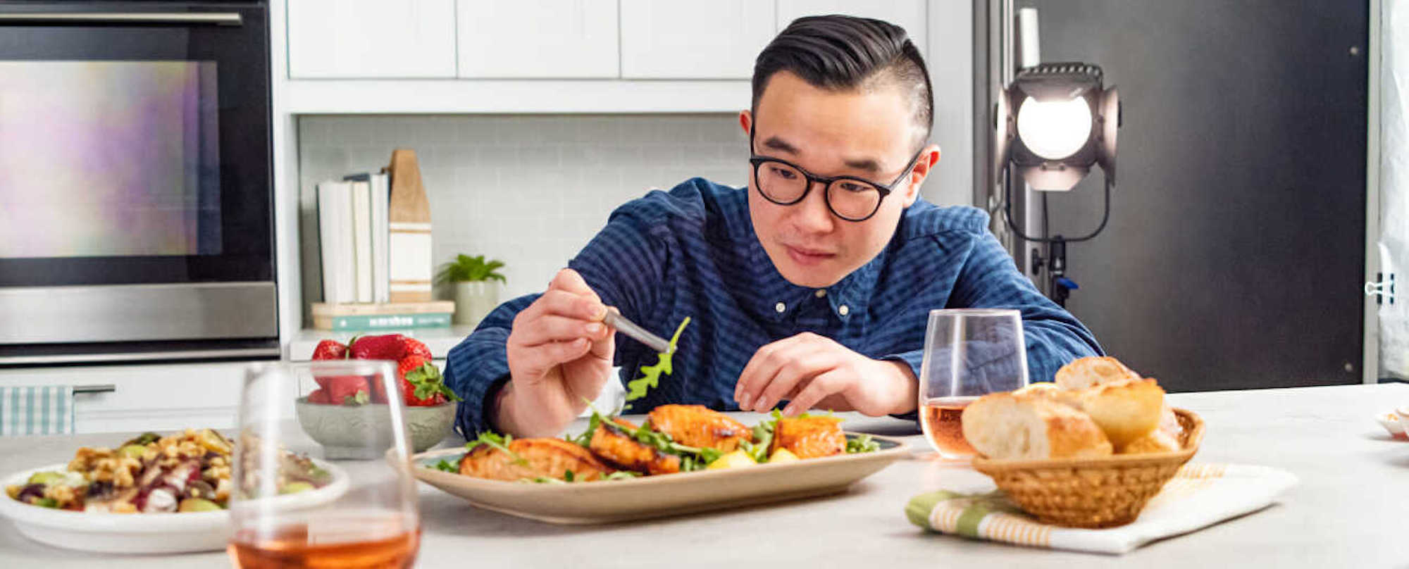 Food stylist arranging greens on a plated meal in a bright kitchen studio with lights and props.