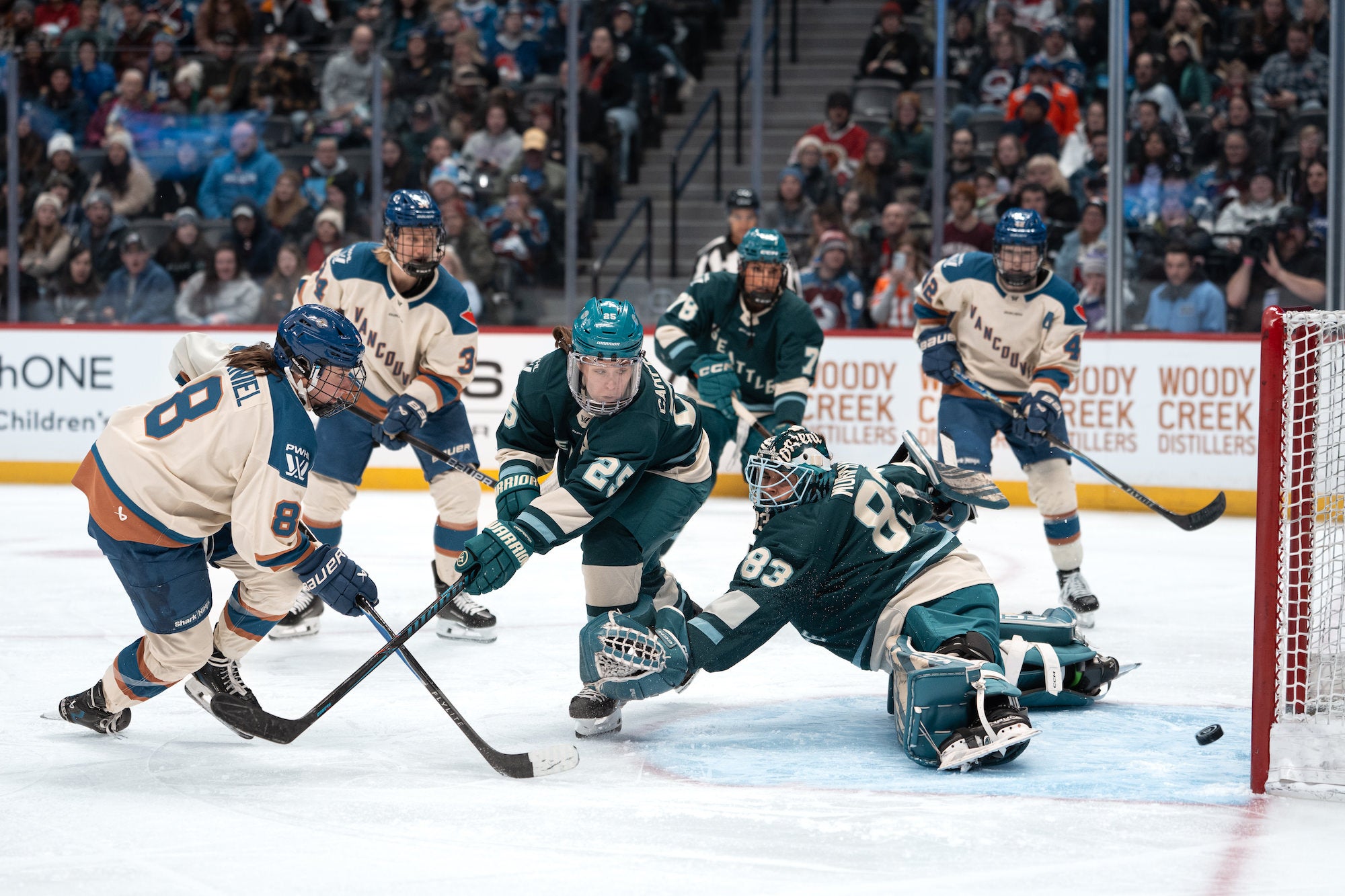 Goalie dives across the crease with glove extended as attackers in cream press near the post.