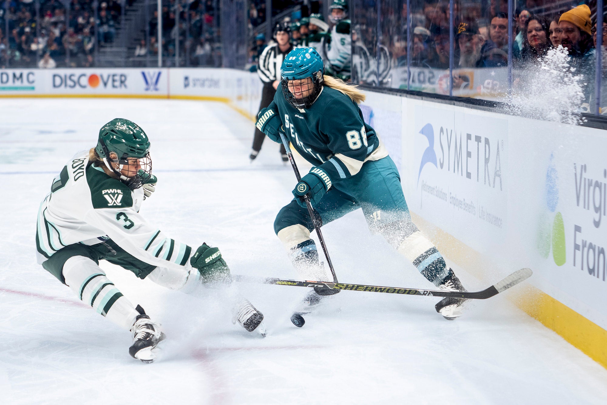 Teal skater shields the puck along the boards while a defender in white slides, kicking up snow.