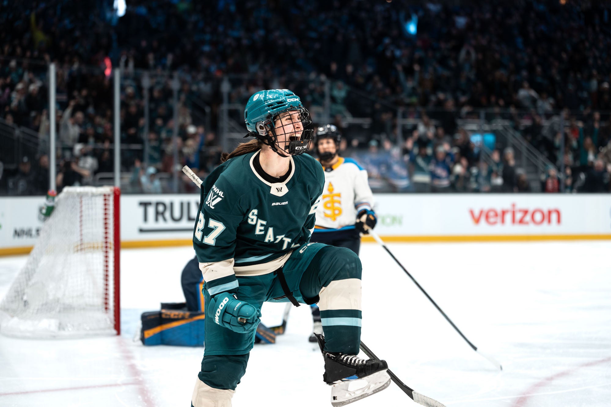 Hockey player in teal jersey celebrates on the ice near the net as a crowd cheers in the background.