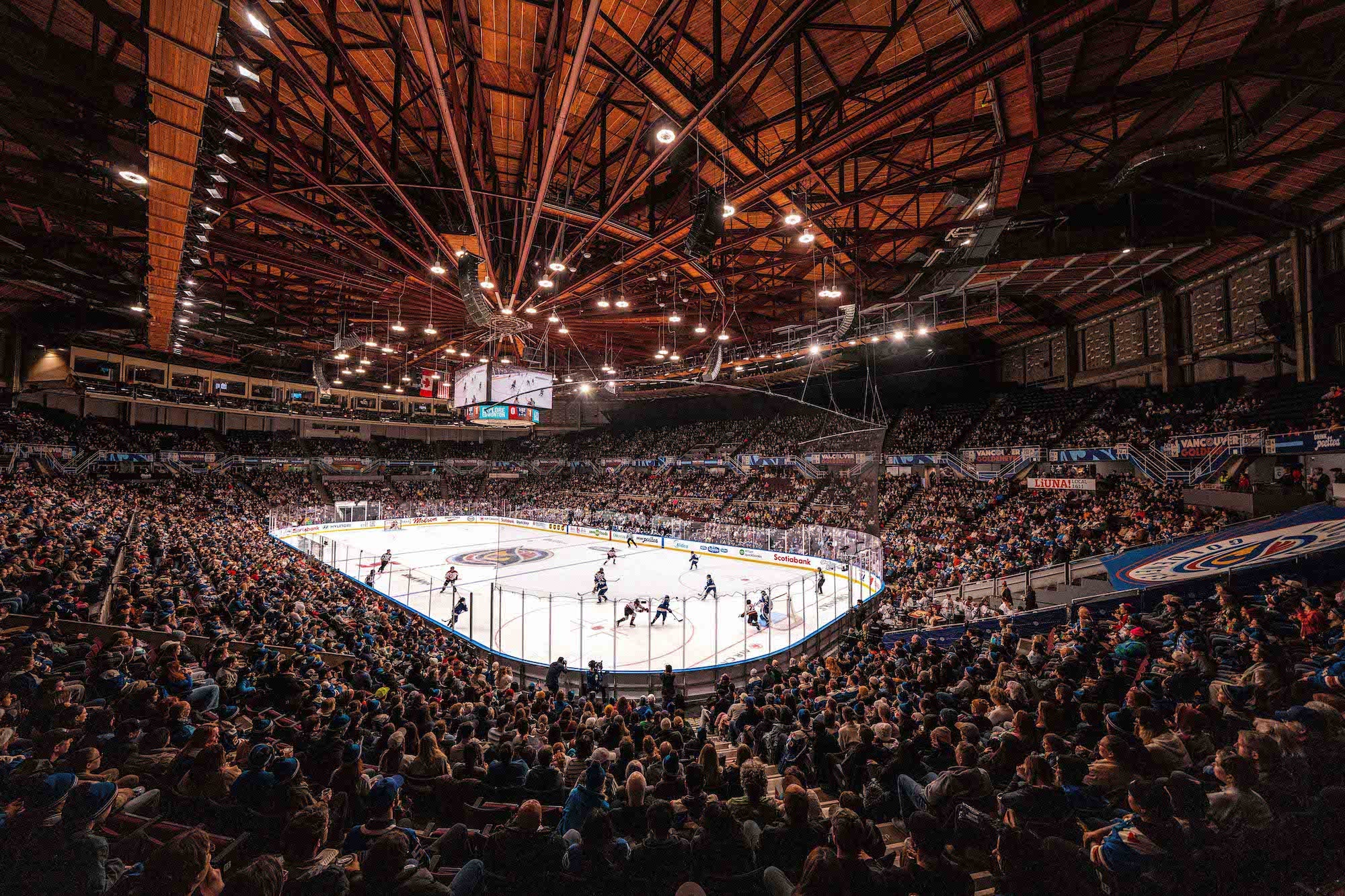 Wide arena view of a sold‑out hockey game under a dramatic wood ceiling and bright rink lights.