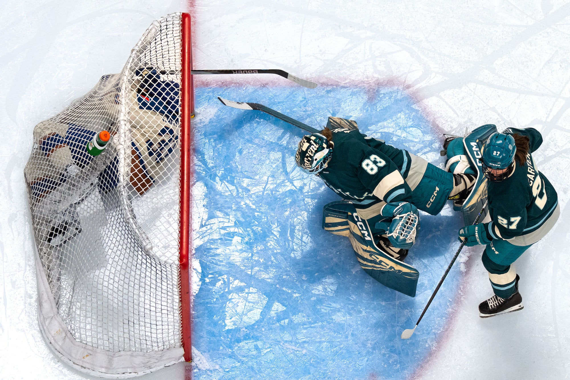Overhead of goalie and defender in teal sweeping a puck away from the goal line at the crease.