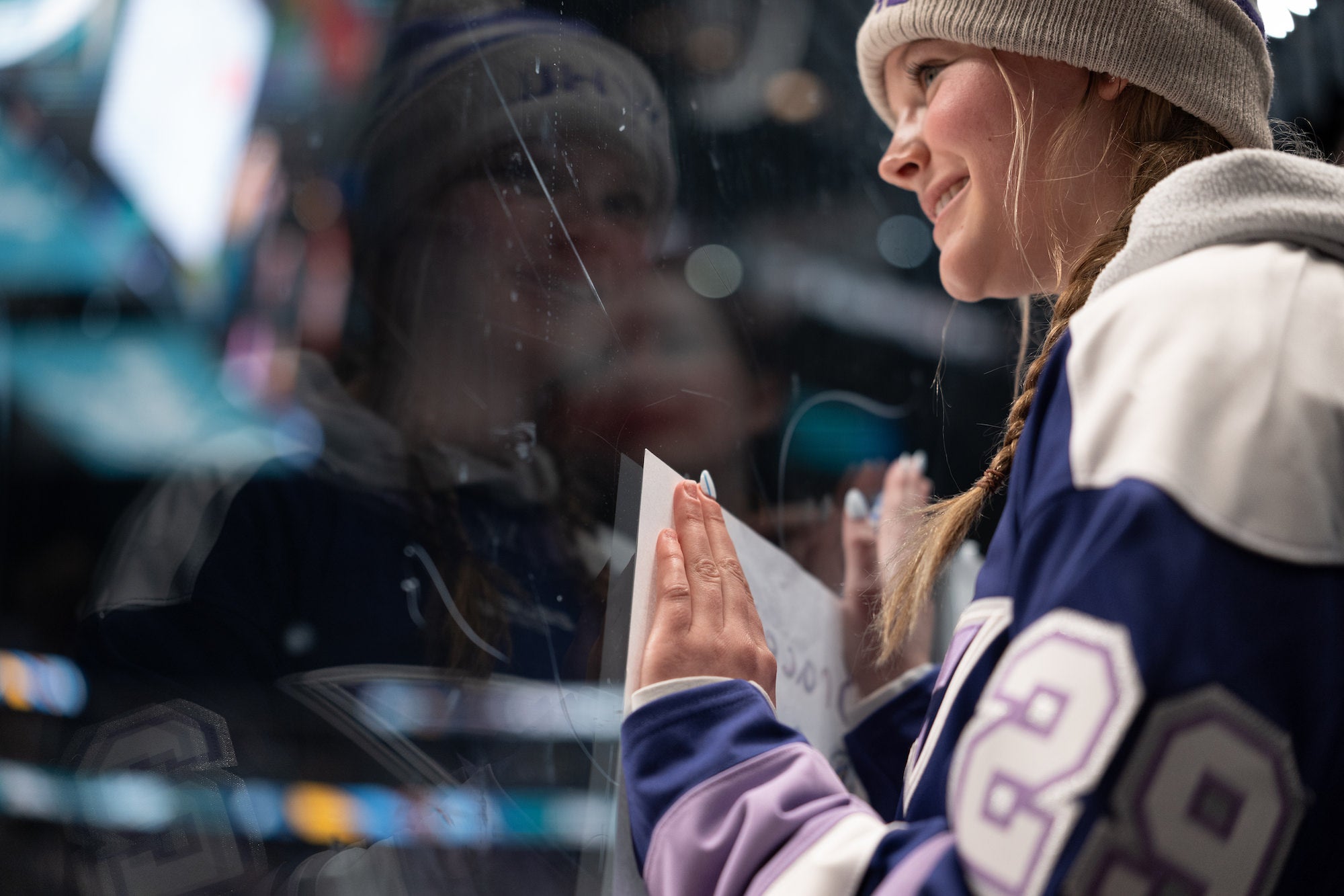 Young fan in a knit hat smiles and presses a handmade sign to the glass at rinkside.