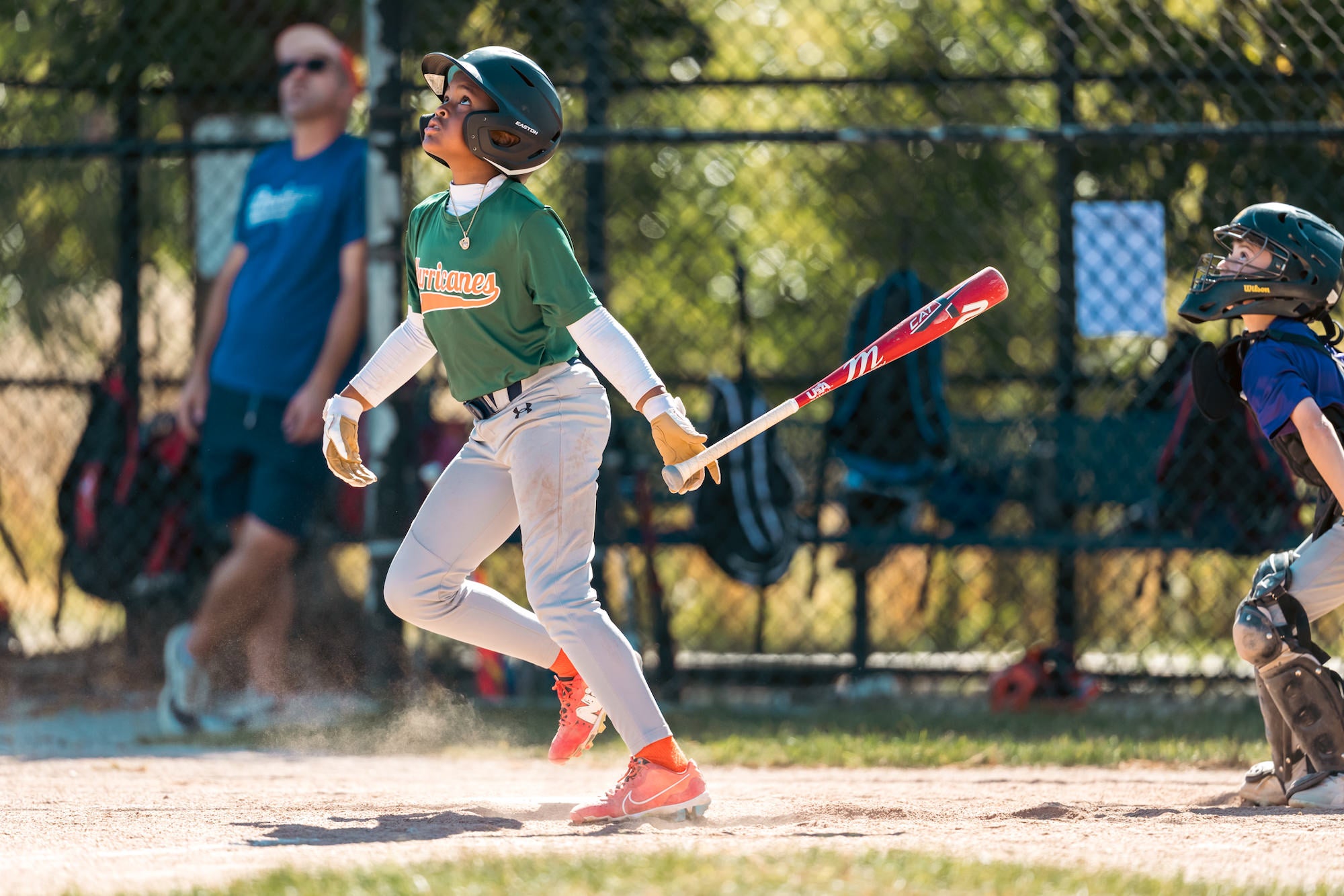 Batter watches a high pop-up near home plate, bat released, catcher and dust visible.