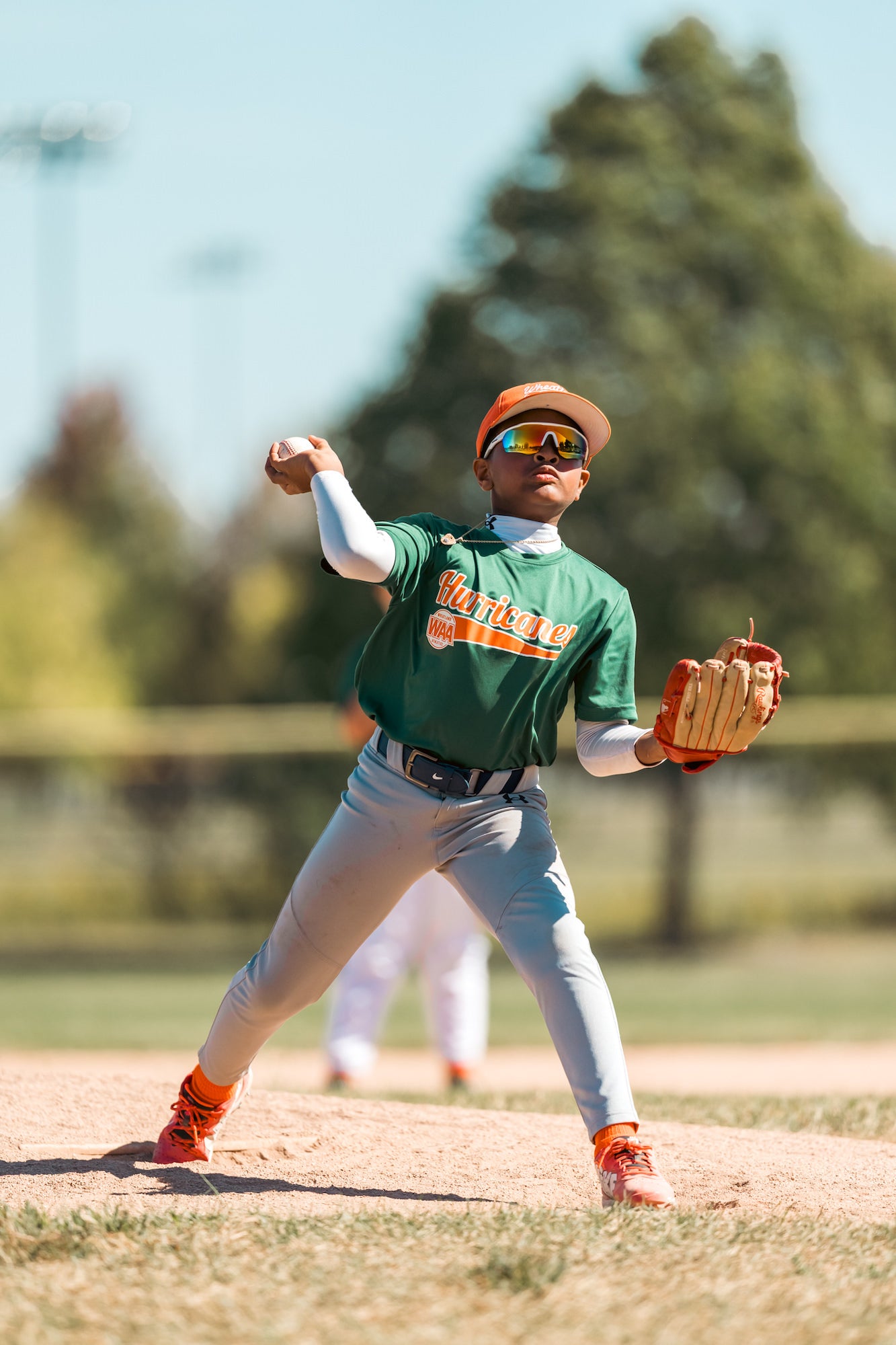Youth pitcher in green uniform fires from the mound, sunglasses gleaming in bright spring light.
