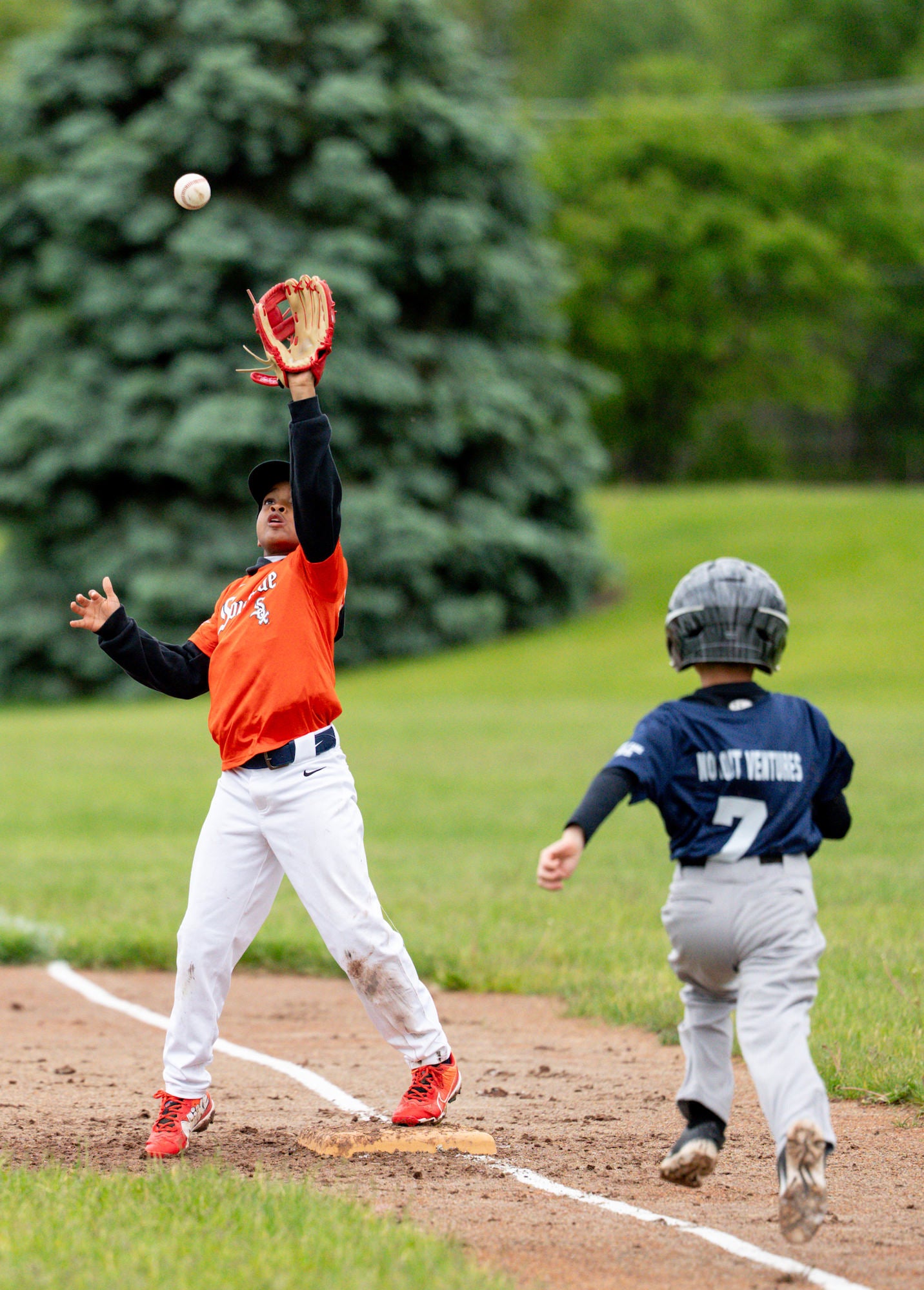 First baseman reaches high for a throw at the bag as the runner charges down the line.