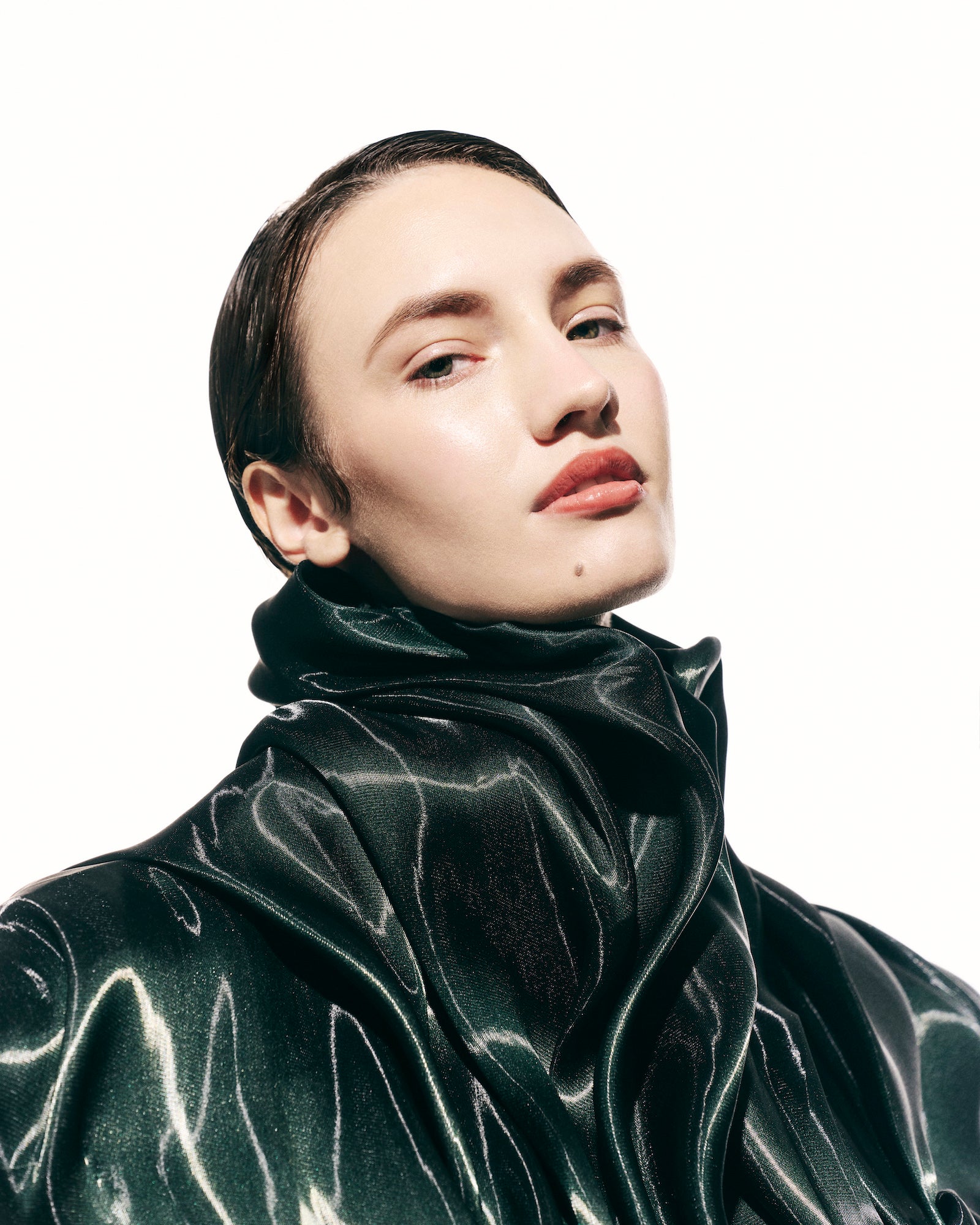 Fashion beauty portrait of a model with slicked-back hair in a high-necked glossy dark green garment against white. Fashion beauty portrait of a model with slicked-back hair in a high-necked glossy dark green garment against white.