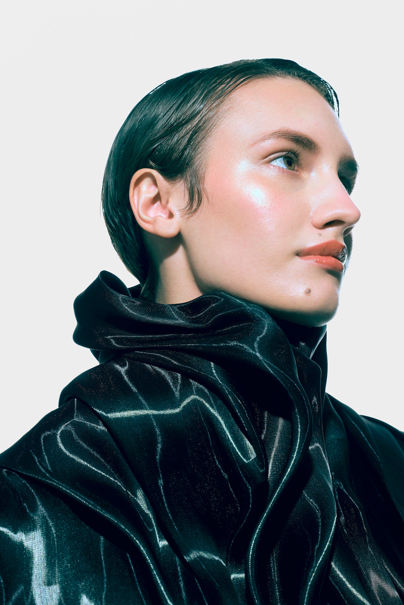 Beauty portrait of a model with slicked-back hair in a glossy black high-collared garment, looking off camera against white. Beauty portrait of a model with slicked-back hair in a glossy black high-collared garment, looking off camera against white.