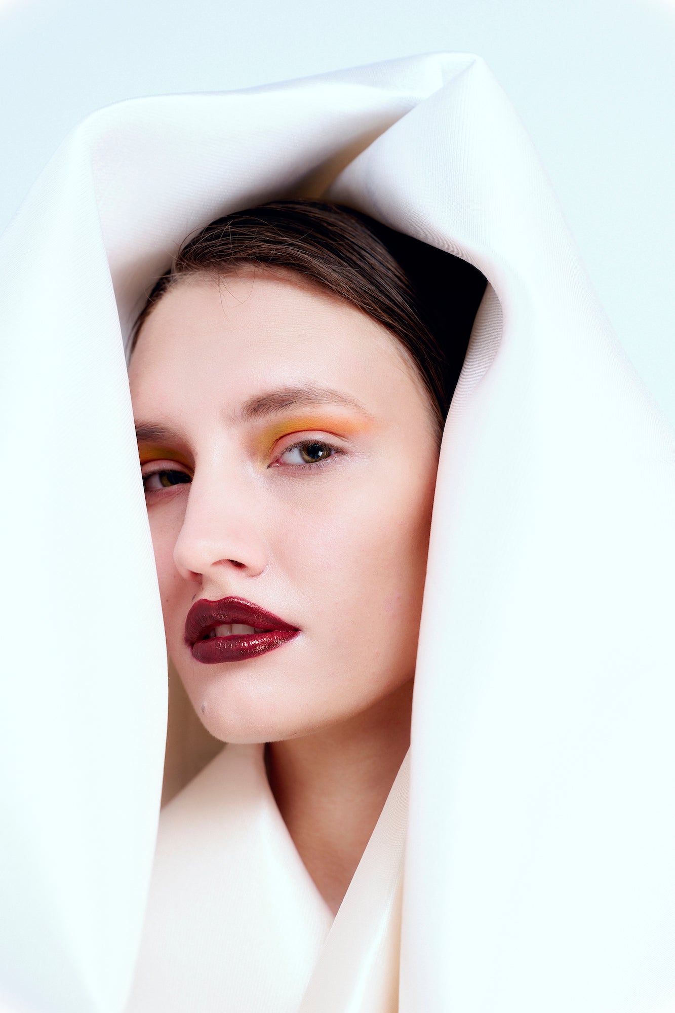 Beauty portrait of a model with orange eyeshadow and dark red lips framed by folds of white fabric. Beauty portrait of a model with orange eyeshadow and dark red lips framed by folds of white fabric.