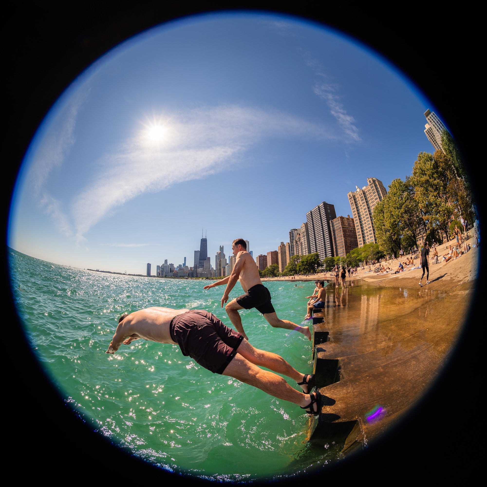 Two people leap from a waterfront ledge into vivid turquoise water as sunbathers line the shore beneath a bright blue sky, captured with a dramatic fisheye view. Photo by Mike Meyers. Alpha 7R V. 8&ndash;14mm f/3.5 Fisheye G. 1/800-sec. f/5.6. ISO 100.