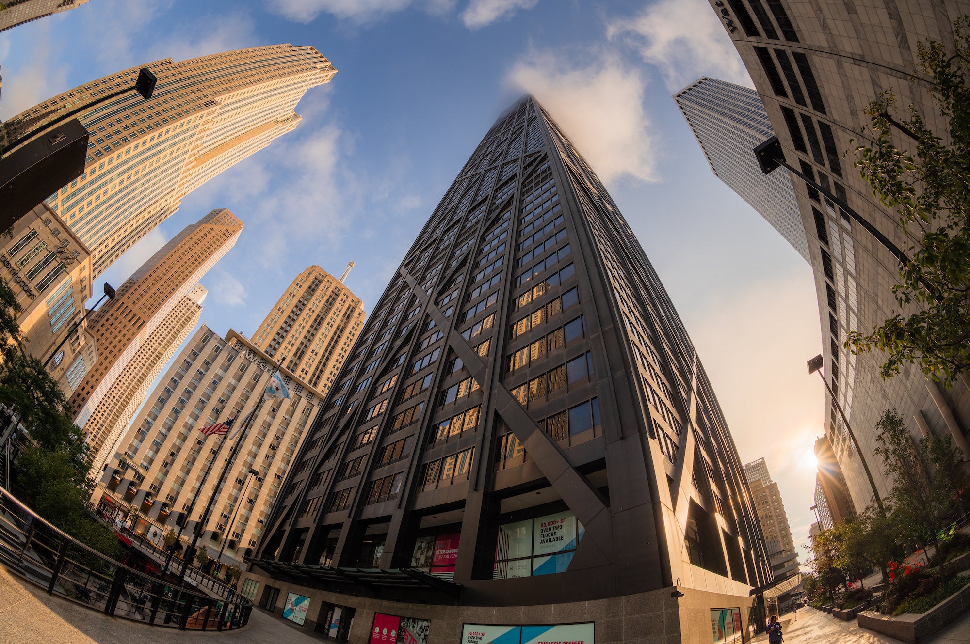 Fisheye view looking up at a cluster of modern high-rise buildings, their curved lines framing a blue sky and low sun. Photo by Mike Meyers. Alpha 7R V. 8-14mm f/3.5 Fisheye G. 1/5000-sec. f/6.3. ISO 100.