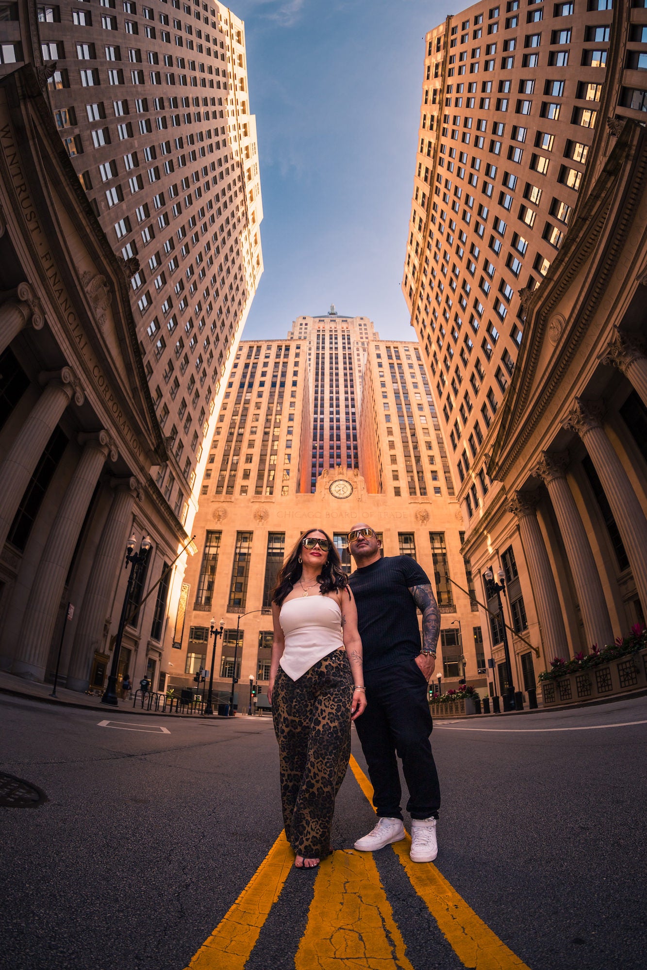 Low-angle fisheye portrait of two people standing on a city street between tall stone office buildings, with a central clock tower rising behind them. Photo by Mike Meyers. Alpha 1. 8-14mm f/3.5 Fisheye G. 1/400-sec. f/3.5. ISO 100.
