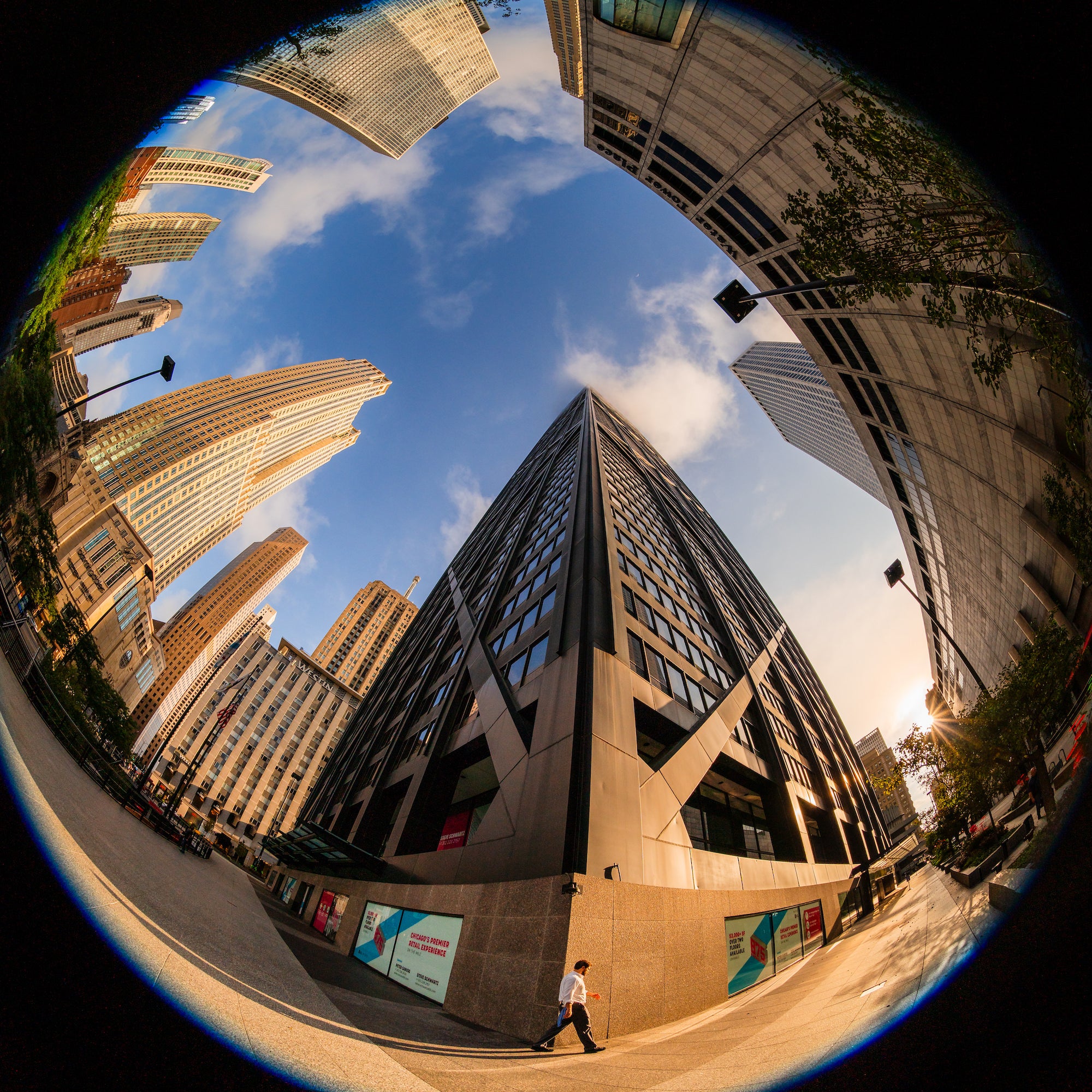 Fisheye view of a pedestrian walking beside modern high-rises, with sunlight and blue sky framed by curved buildings. Photo by Mike Meyers. Alpha 7R V. 8-14mm f/3.5 Fisheye G. 1/1250-sec. f/6.3. ISO 100.