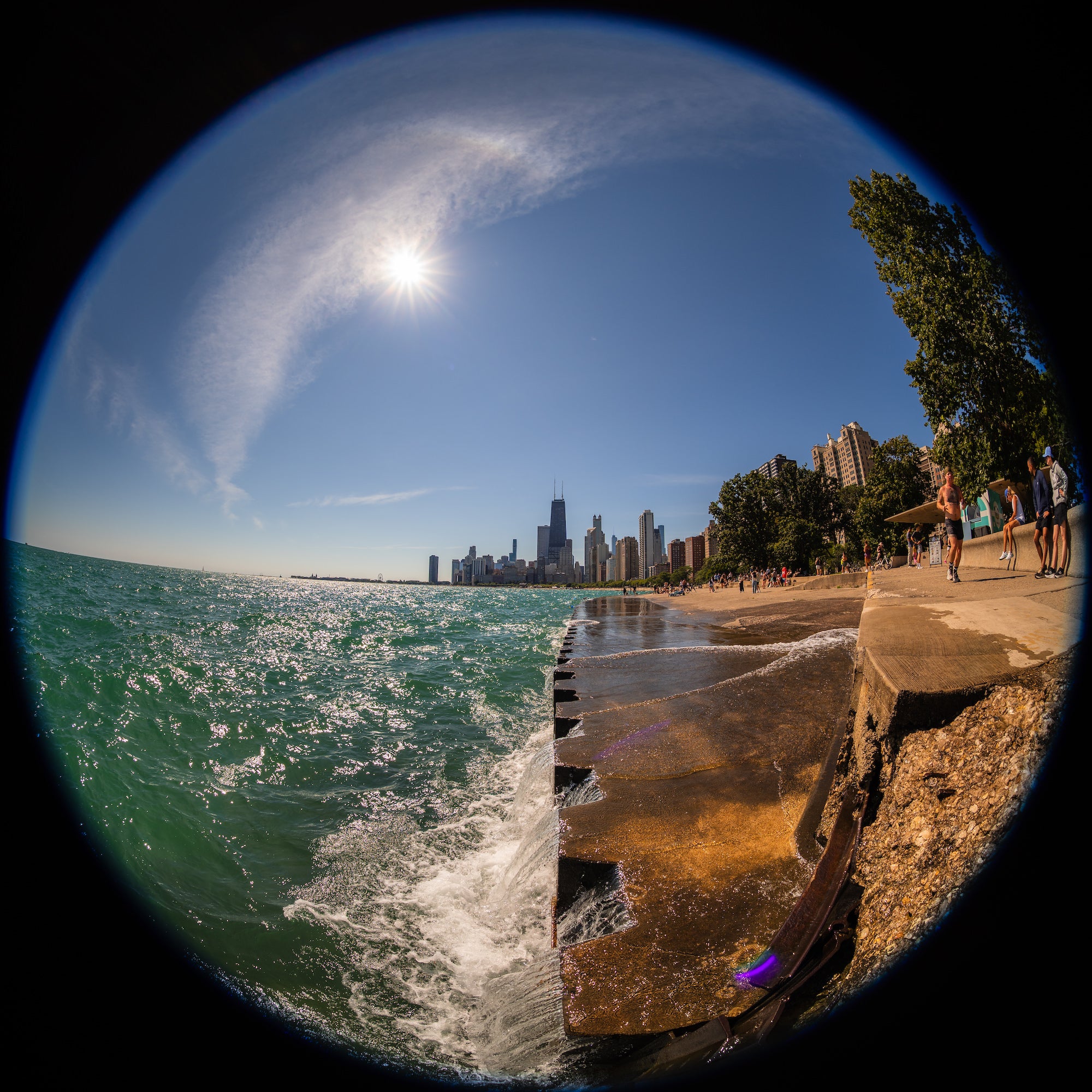 Fisheye view of sunlit waves breaking along a concrete shoreline, with people on the waterfront promenade and tall buildings rising in the distance. Photo by Mike Meyers. Alpha 7R V. 8-14mm f/3.5 Fisheye G. 1/800-sec. f/5.6. ISO 100.