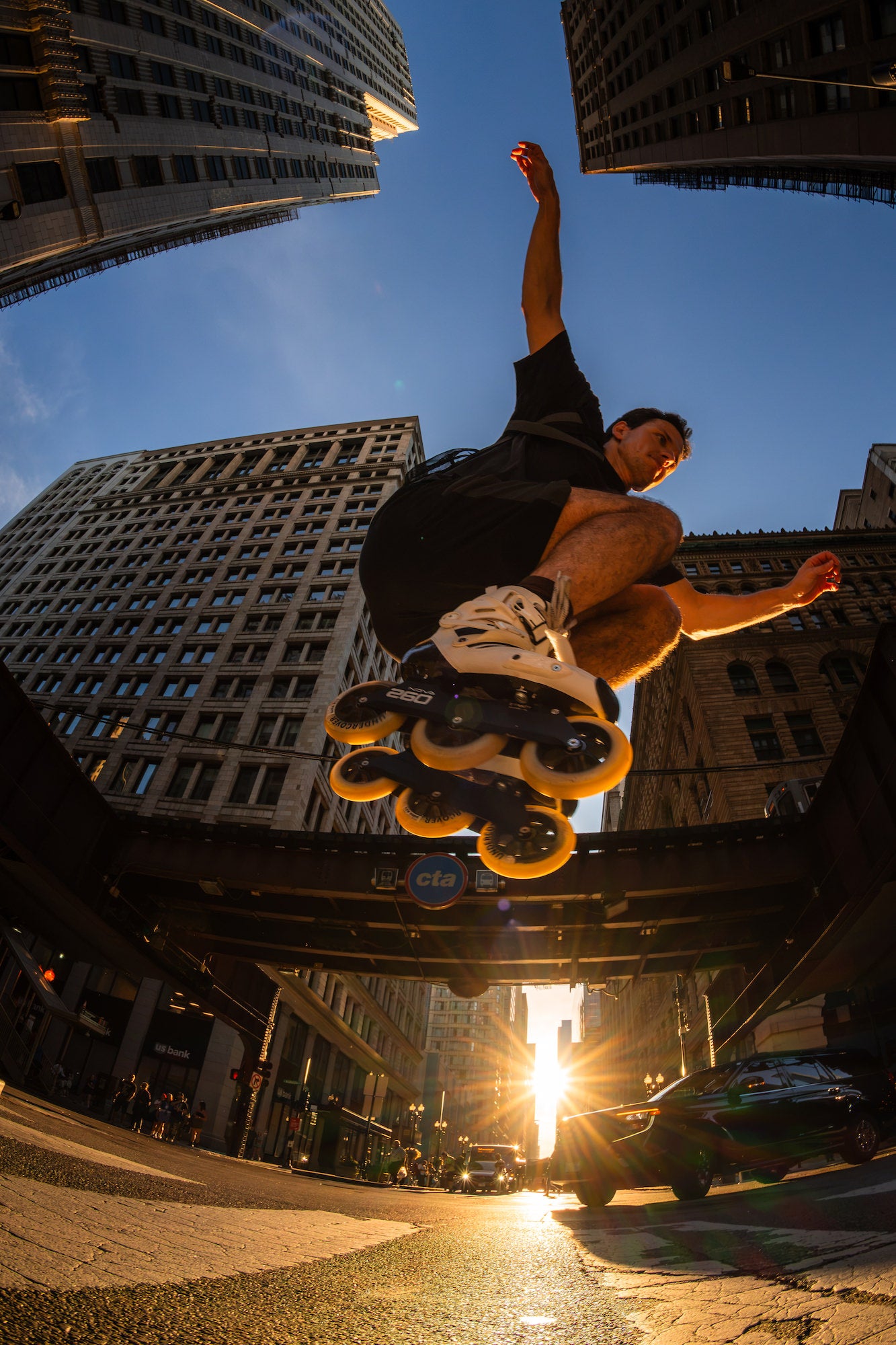 Low-angle fisheye view of an inline skater jumping above a sunlit downtown street and elevated train tracks. Photo by Mike Meyers. Alpha 1. 8-14mm f/3.5 Fisheye G. 1/1000-sec. f/10. ISO 500.