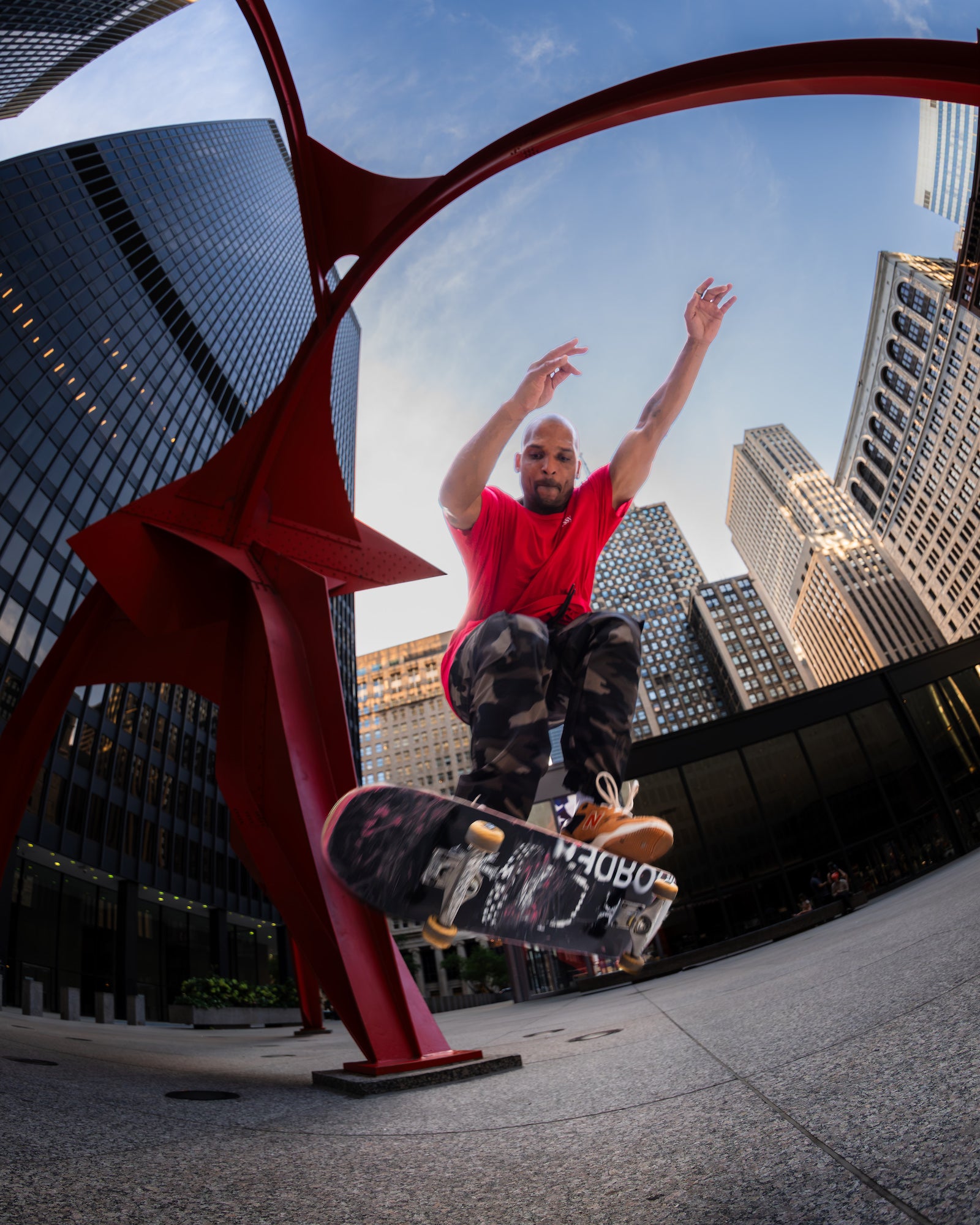 Low-angle fisheye view of a skateboarder midair beside a red steel sculpture, framed by modern high-rise buildings. Photo by Mike Meyers. Alpha 1. 8-14mm f/3.5 Fisheye G. 1/500-sec. f/3.5. ISO 500.