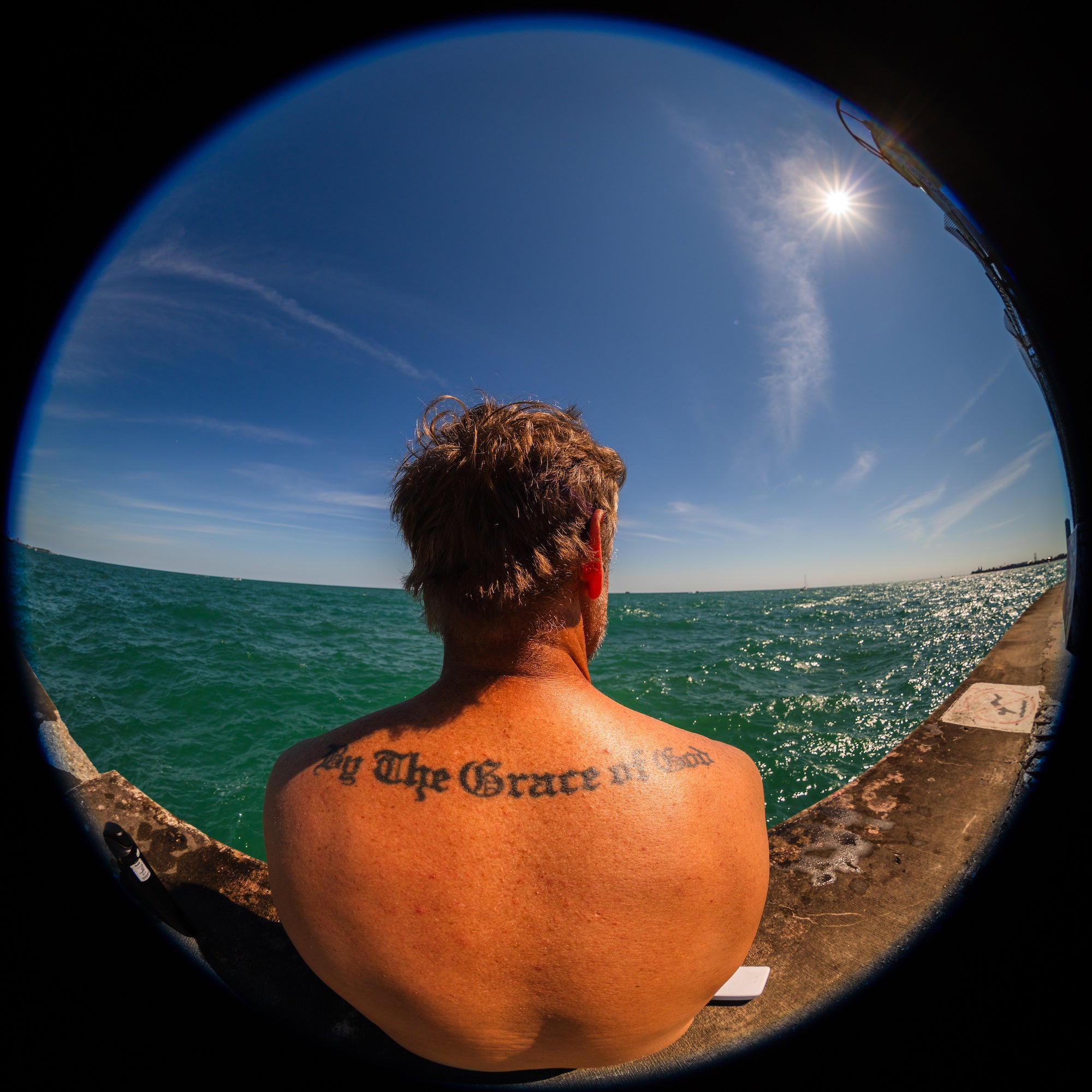 A shirtless man with a shoulder tattoo sits on a concrete pier facing choppy turquoise water beneath a bright sun, captured with a dramatic fisheye view. Photo by Mike Meyers. Alpha 7R V. 8&ndash;14mm f/3.5 Fisheye G. 1/1600-sec. f/5.6. ISO 100.