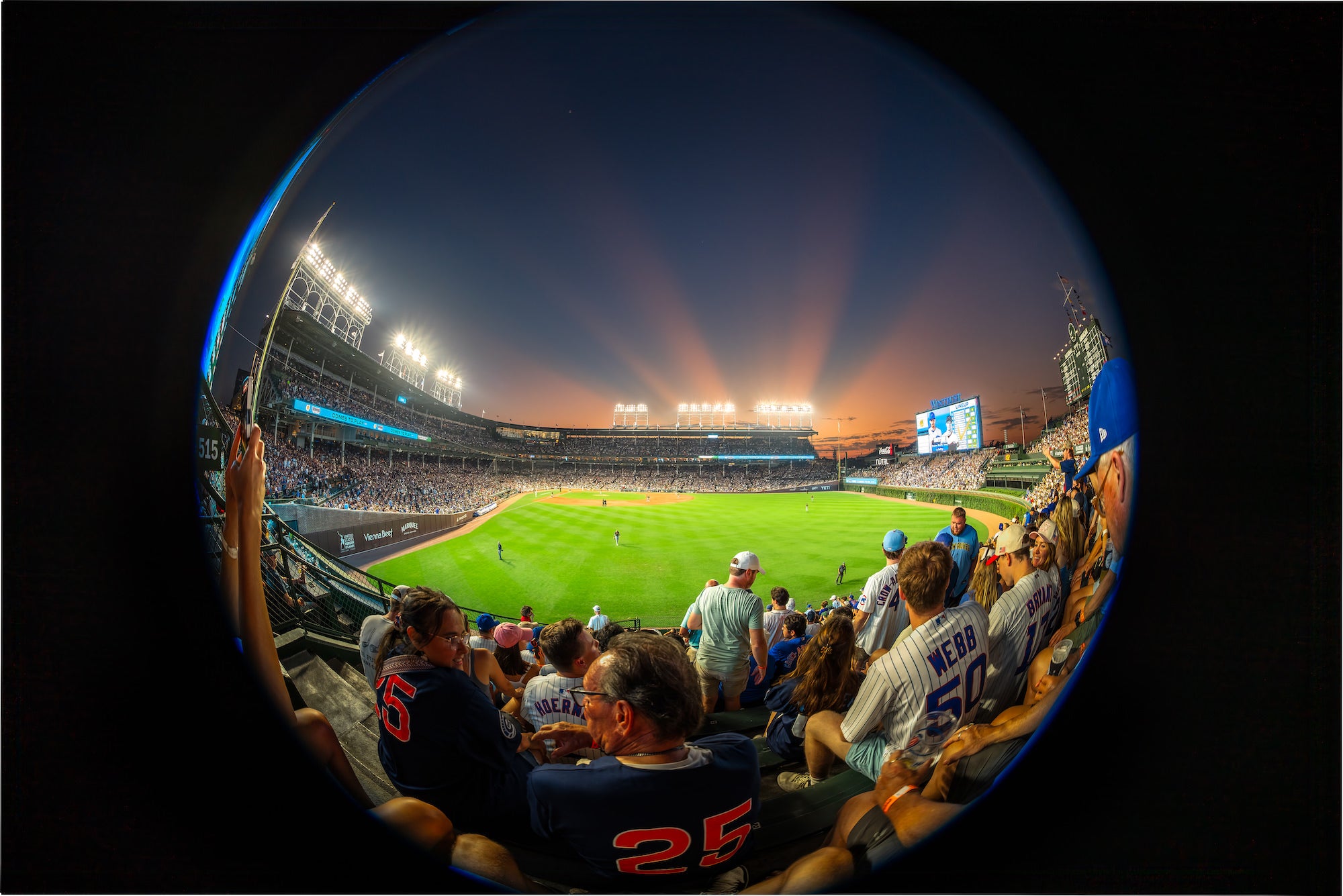 A packed baseball stadium glows beneath bright field lights and a colorful sunset sky, captured from the grandstands with a dramatic fisheye view. Photo by Mike Meyers. Alpha 7R V. 8&ndash;14mm f/3.5 Fisheye G. 1/200-sec. f/4. ISO 320.