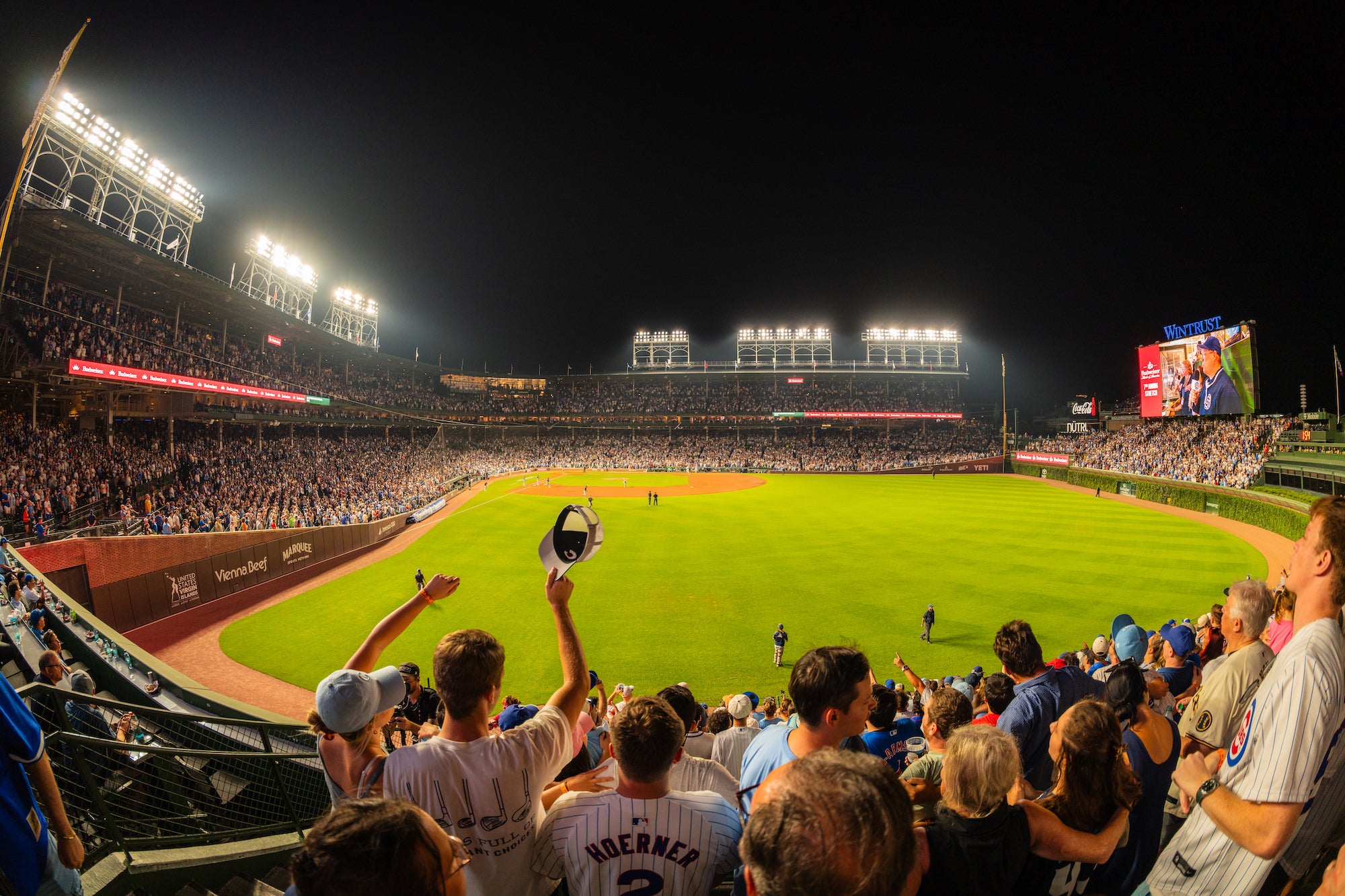 Fans fill a brightly lit baseball stadium at night, cheering from grandstands around the green field; one raises a cap in the foreground. Photo by Mike Meyers. Alpha 7R V. 8-14mm f/3.5 Fisheye G. 1/200-sec. f/3.5. ISO 500.