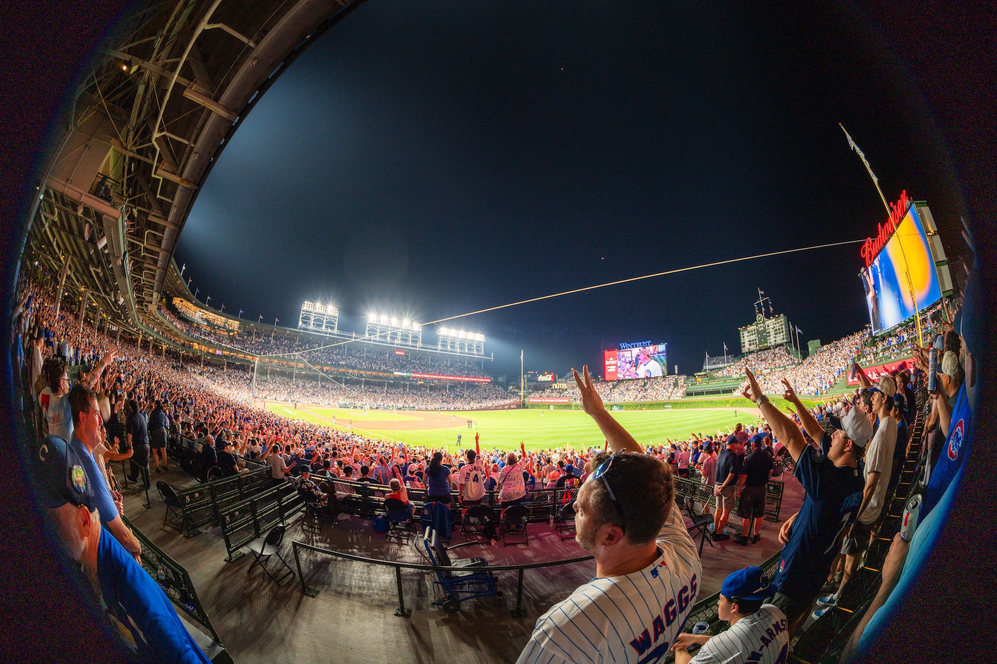Fans cheer with raised arms during a nighttime baseball game, viewed through a fisheye lens from the stadium stands. Photo by Mike Meyers. Alpha 7R V. 8-14mm f/3.5 Fisheye G. 1/80-sec. f/3.5. ISO 320.