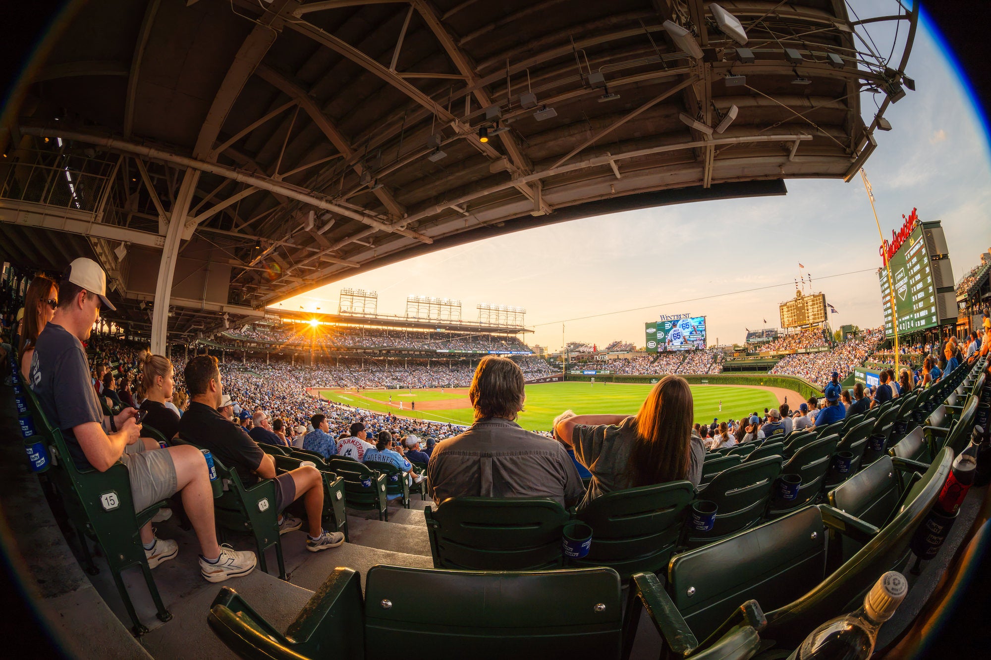 Fans watch a baseball game from covered stadium seats as the setting sun flares beyond the grandstand, illuminating the field and packed bleachers. Photo by Mike Meyers. Alpha 7R V. 8-14mm f/3.5 Fisheye G. 1/80-sec. f/16. ISO 100.