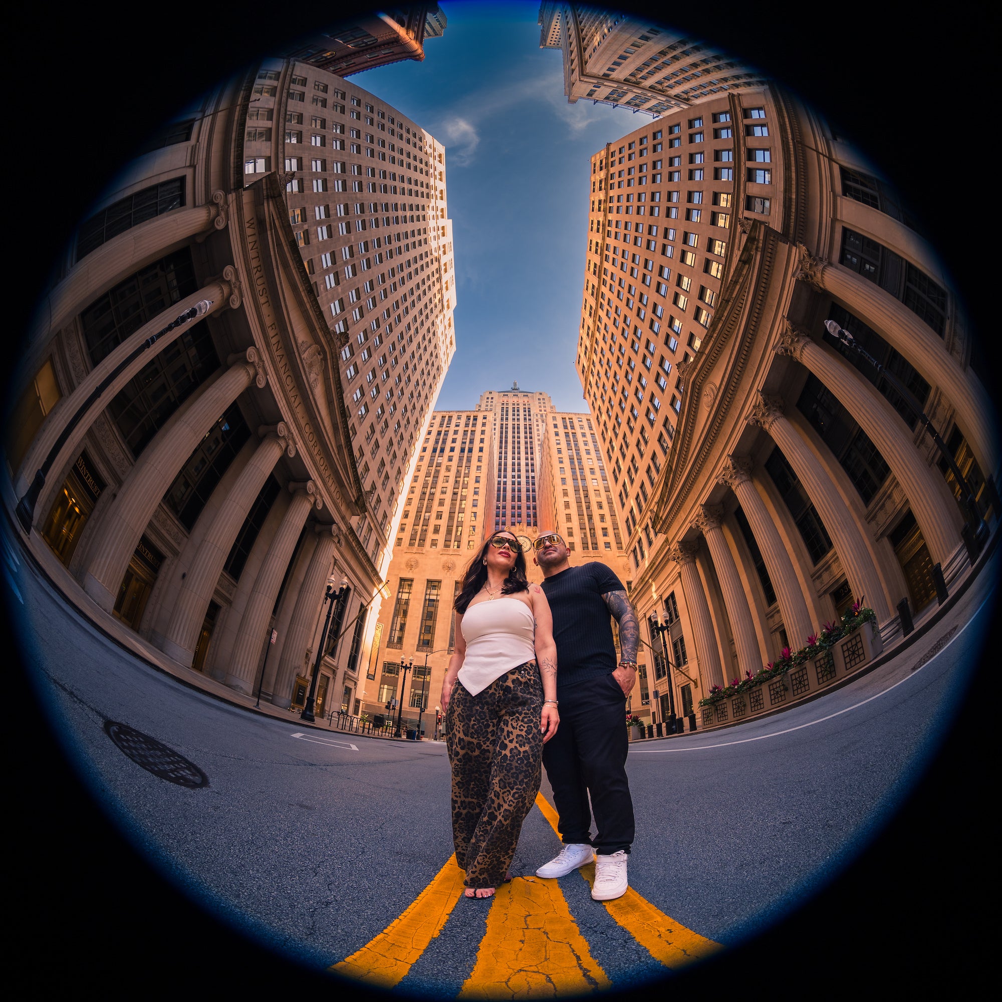 Fisheye portrait of two people standing on a yellow-striped city street, framed by tall stone buildings and a central clock tower. Photo by Mike Meyers. Alpha 1. 8-14mm f/3.5 Fisheye G. 1/100-sec. f/3.5. ISO 100.