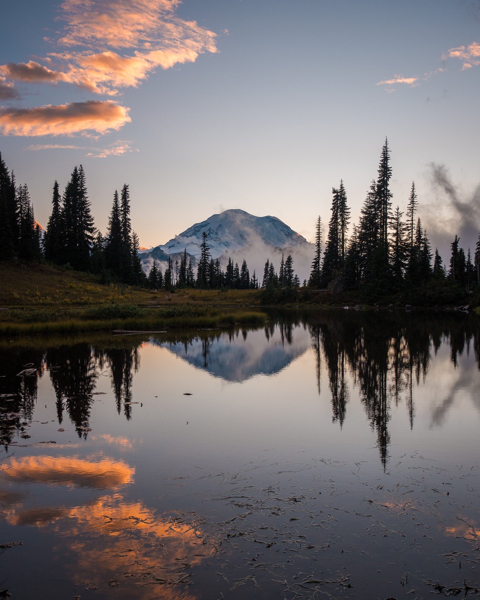 Snow-covered mountain reflected in a still lake surrounded by evergreen trees at sunset. Photographed by Nate Luebbe.