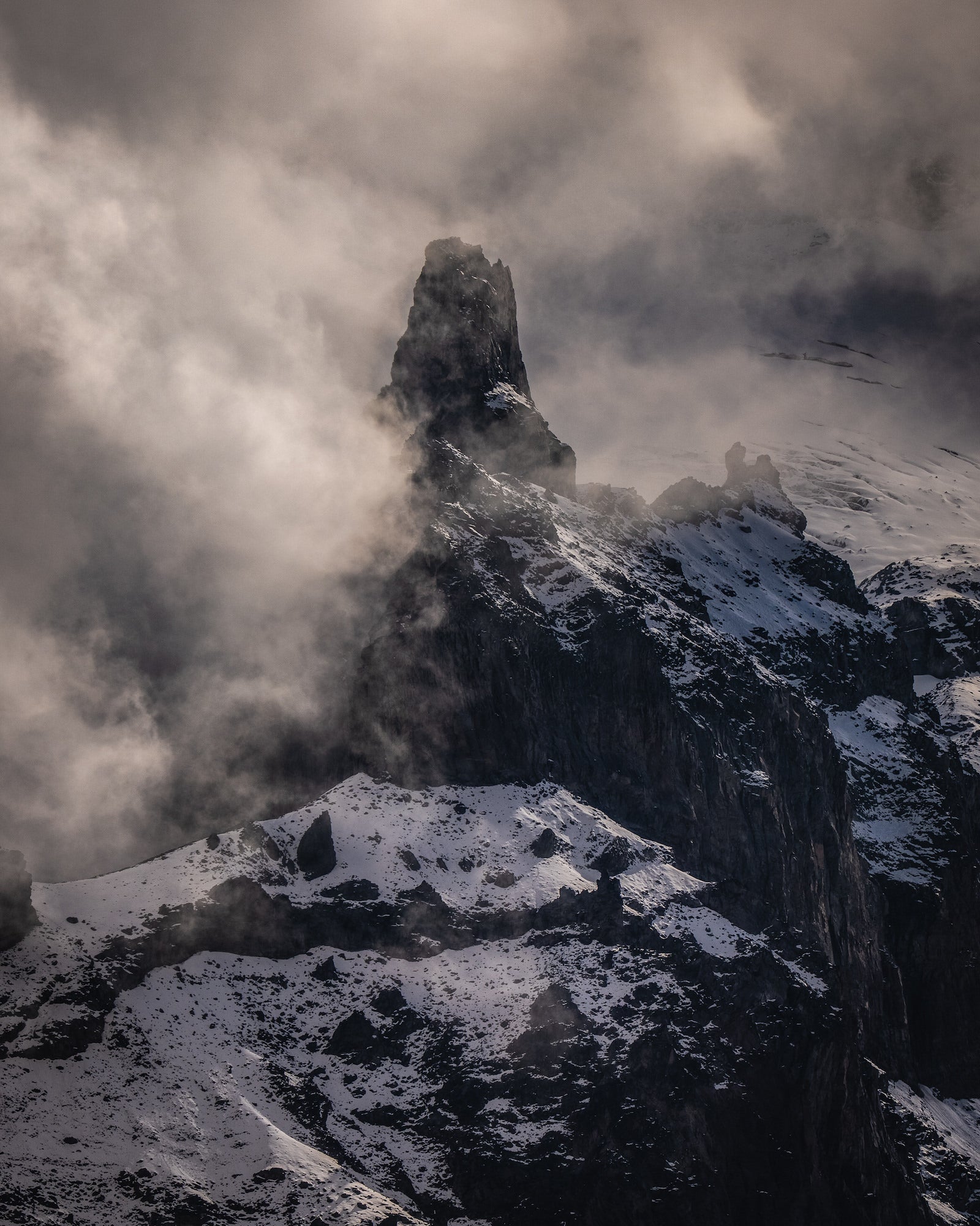 A jagged, snow-dusted mountain spire rises through thick swirling clouds, its dark rock face and surrounding ridgelines emerging from the mist. Photo by Nate Luebbe. 400mm f/4.5 G Master OSS.